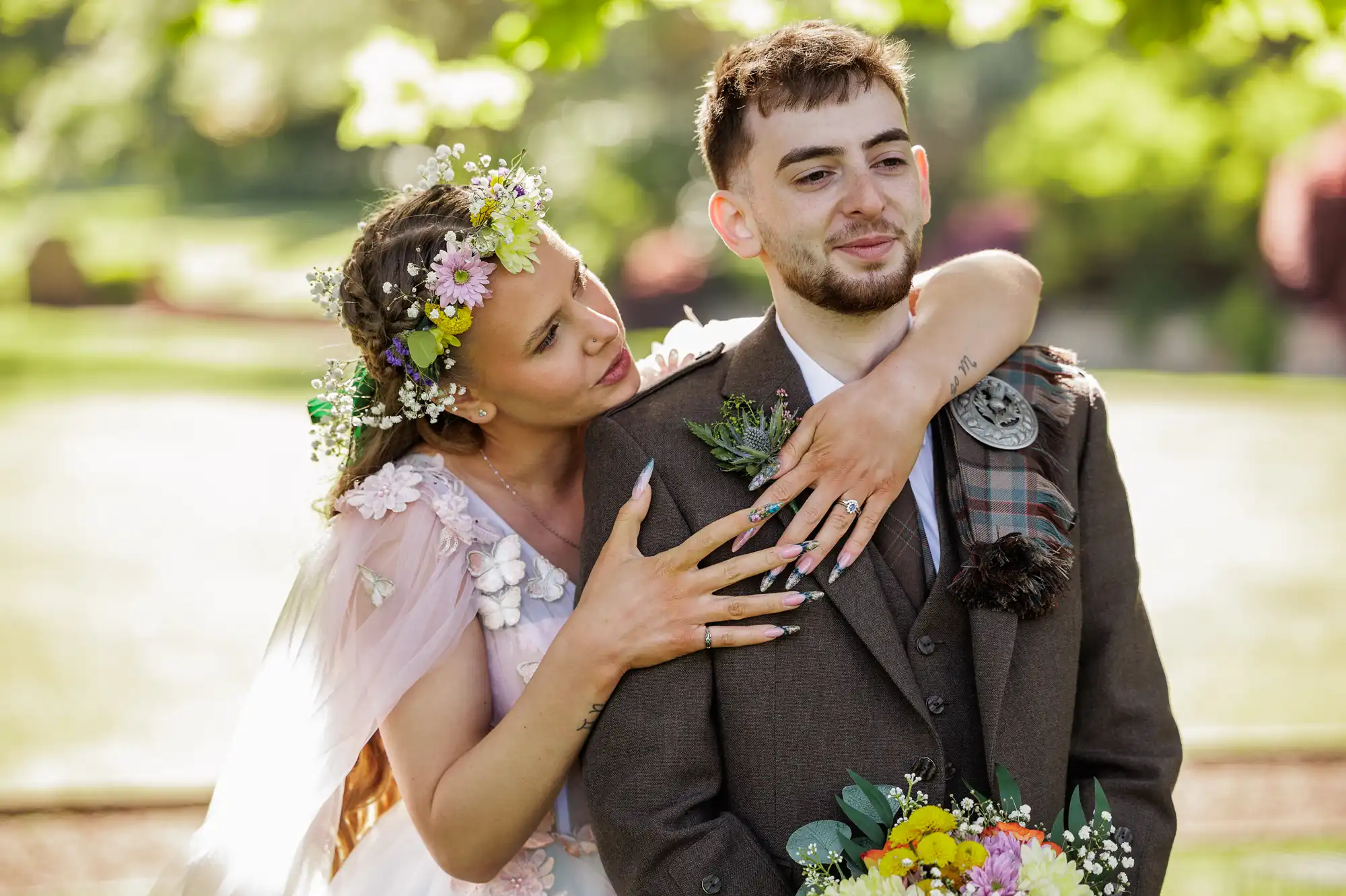 A smiling woman in a floral dress and flower crown hugs a man in a brown suit and tartan sash from behind outdoors. She rests her chin on his shoulder and holds his chest, whilst he smiles, holding a bouquet. Sunlight filters through the green trees.