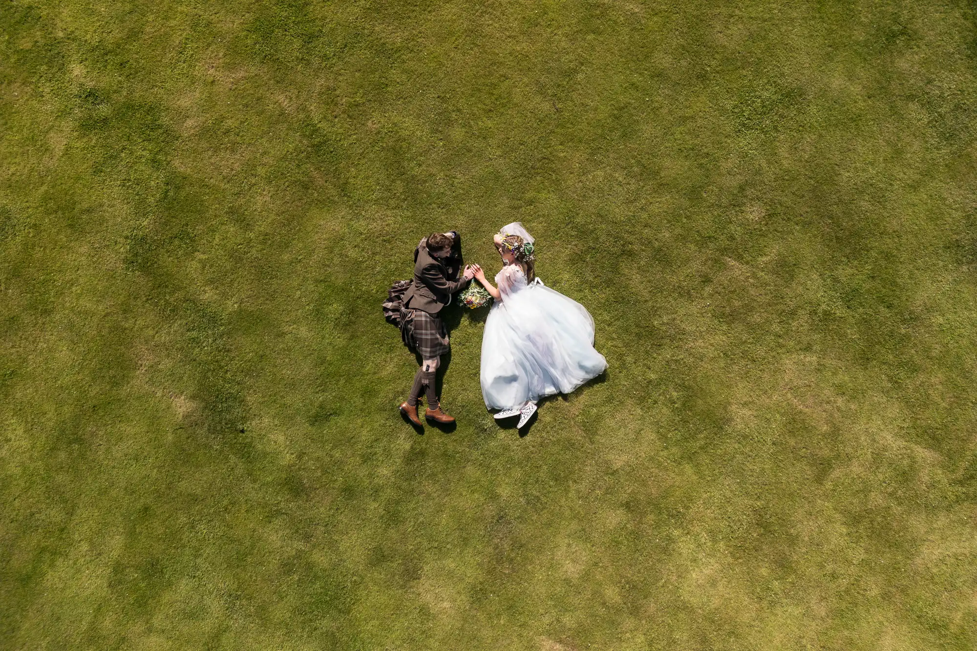 A bride in a white dress and a groom in a dark suit lie side by side on green grass, holding hands and looking at each other. The photo is taken from above, showing them surrounded by the wide expanse of lawn.