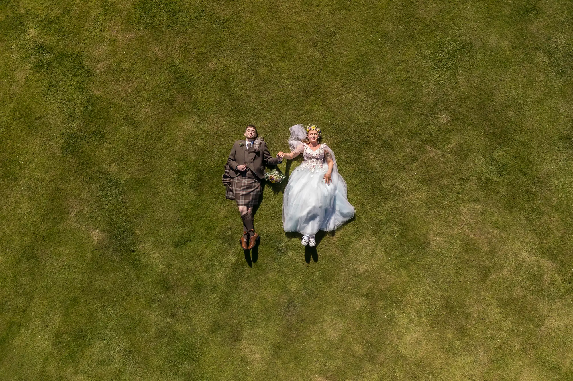 A bride and groom lie side by side on green grass, holding hands. The bride wears a white dress and floral crown, while the groom is in a brown suit and kilt. They look up, smiling, with a bouquet near them, viewed from directly above.