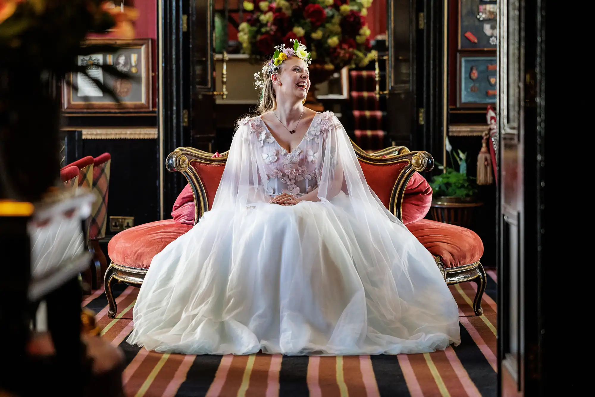 A smiling bride in a white, floral wedding dress and flower crown sits on a vintage red velvet sofa in an ornate, richly decorated room with dark walls, striped carpet, and floral arrangements in the background.