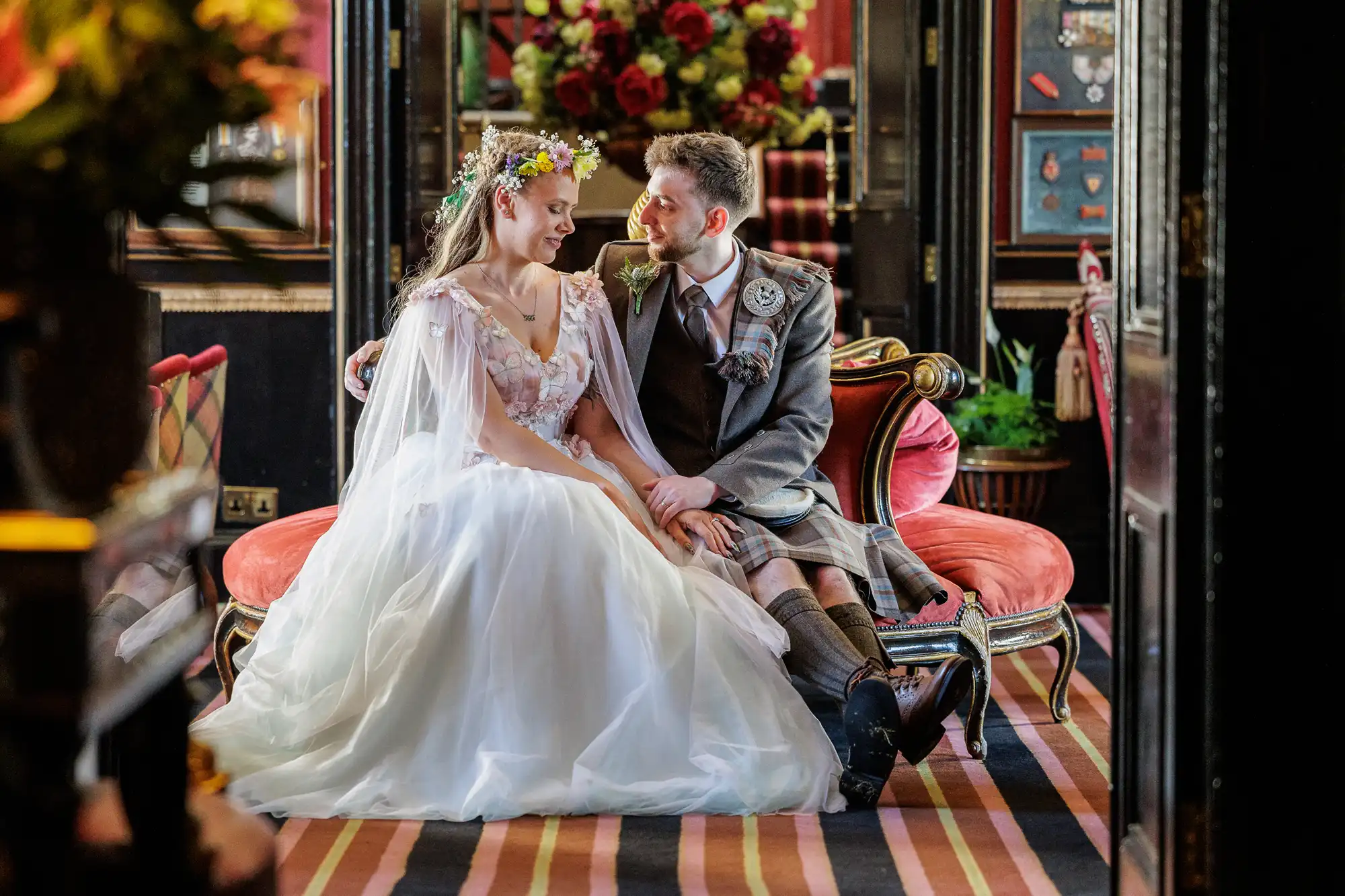 A bride in a white gown and flower crown sits on a vintage red sofa beside a groom in a kilt. They gaze at each other, smiling, holding hands. The setting is richly decorated with striped carpet, dark walls, and a display of red flowers in the background.