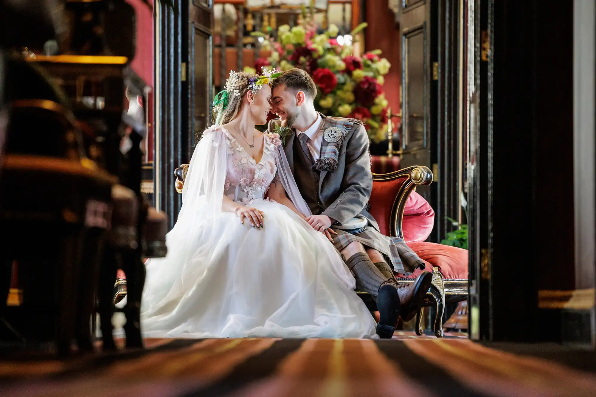 A bride in a white dress and floral crown sits closely with a groom in a grey kilt and jacket on an ornate bench, touching noses and smiling. They are in an elegant, dimly lit room with flowers and dark wood, creating a romantic atmosphere.