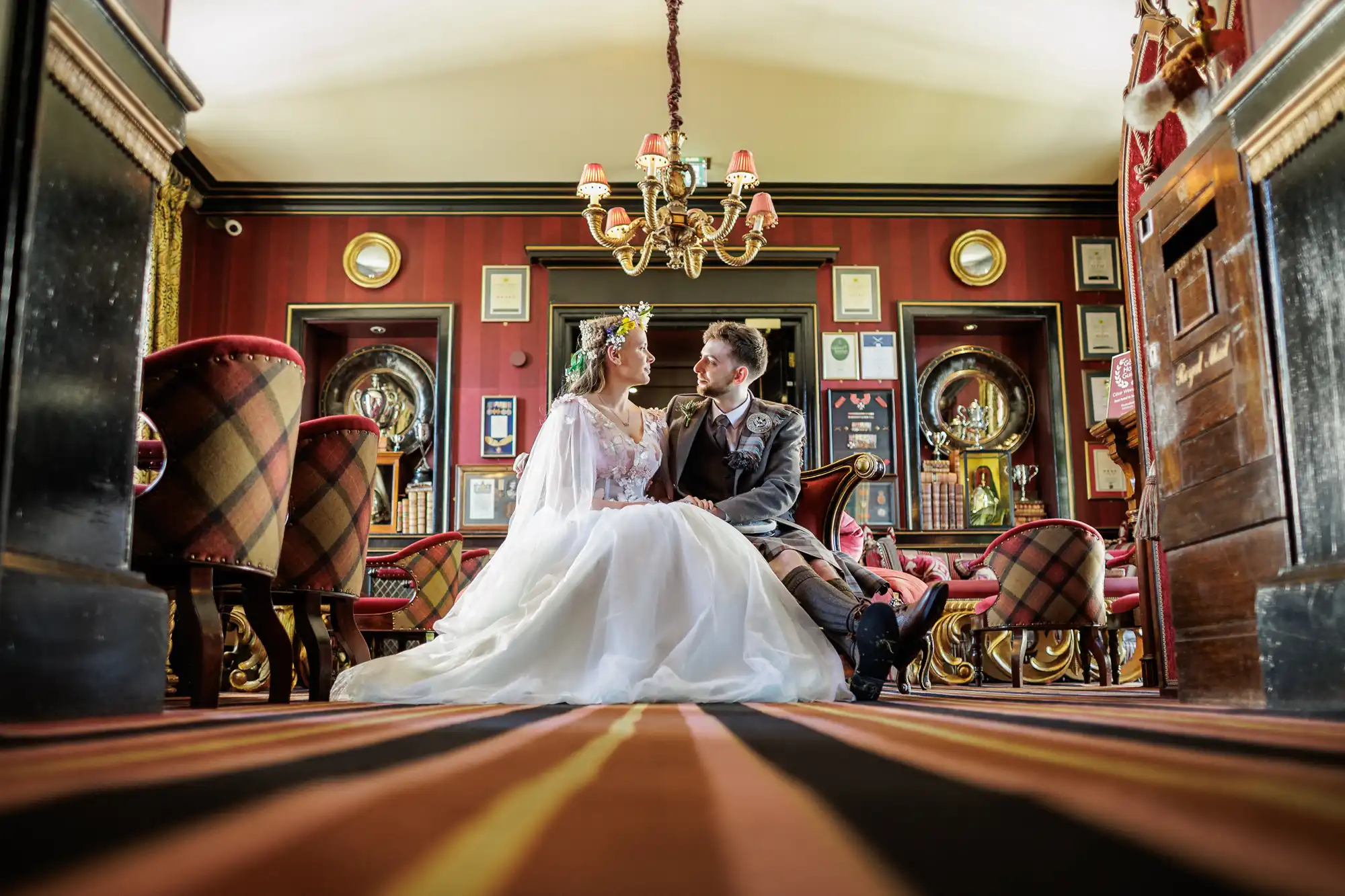 A bride in a flowing white gown and a groom in a formal suit sit closely together on a striped carpet in an ornate, richly decorated room. They gaze at each other, surrounded by vintage decor, red walls, framed pictures, and a chandelier above.