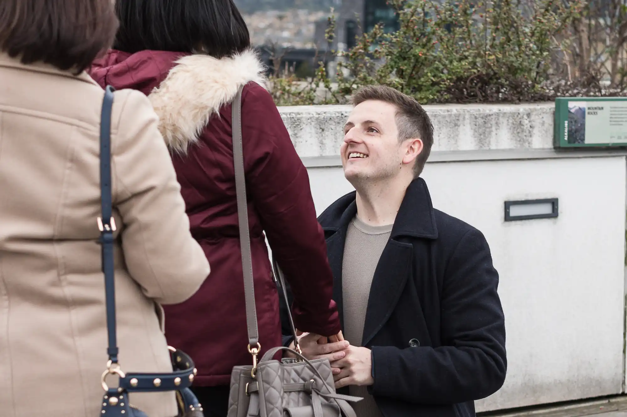 A man smiling and looking up, standing outdoors in conversation with two women who are facing away from the camera.