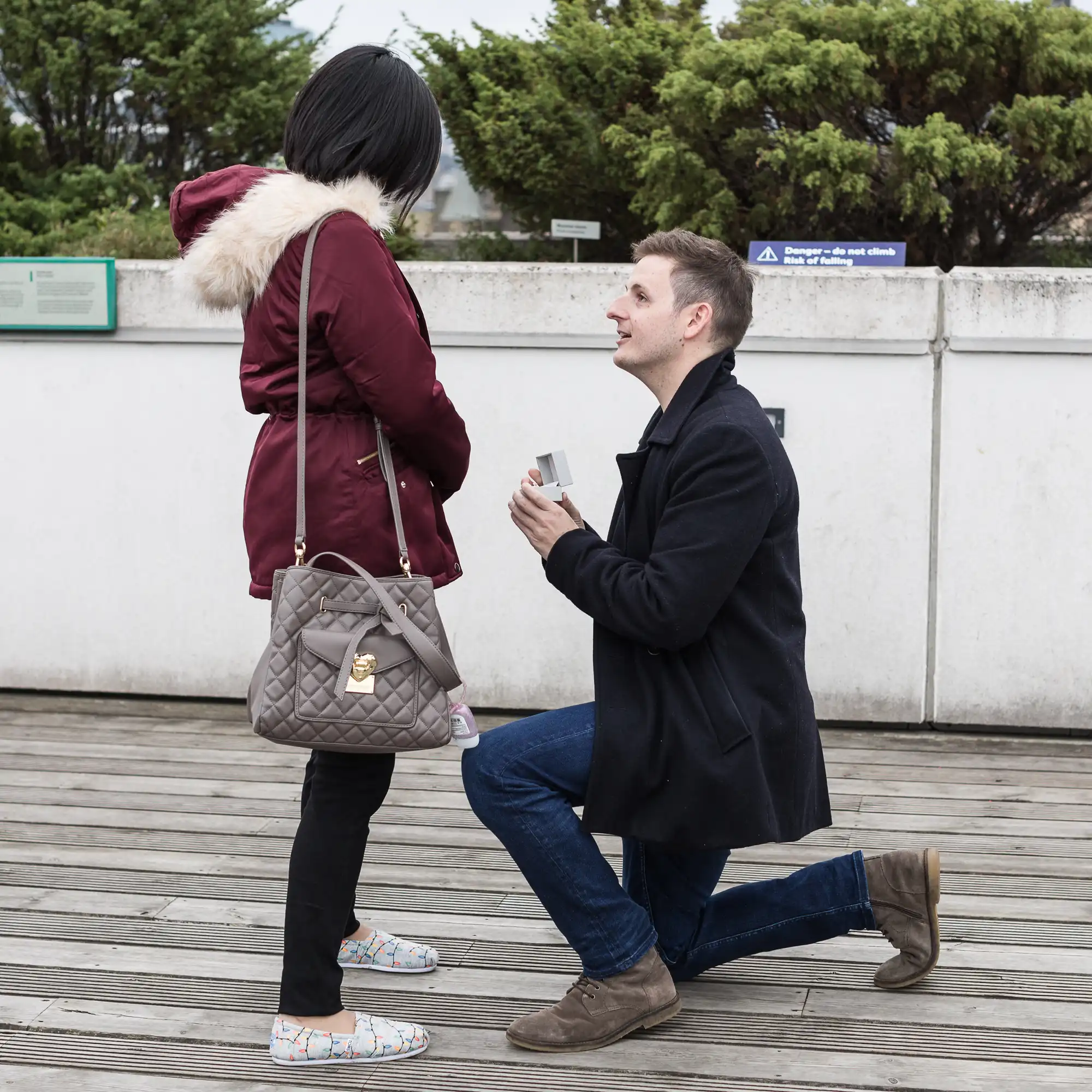 Man kneeling and holding a ring box in front of a woman on an outdoor balcony, appearing to propose.