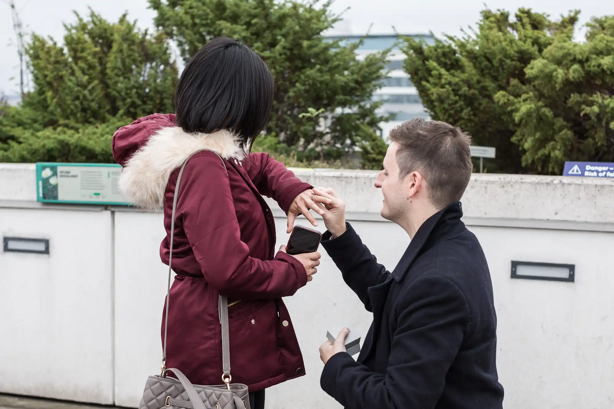 Man proposing to woman on one knee, holding her hand, outdoors with greenery and a building in the background.