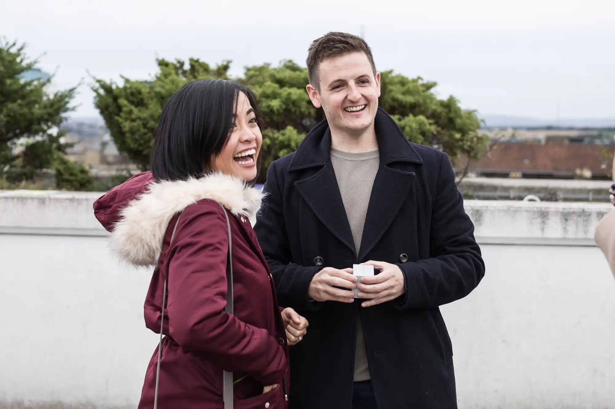 Two people smiling and talking outdoors, a woman in a red jacket and a man in a dark coat, holding coffee cups.