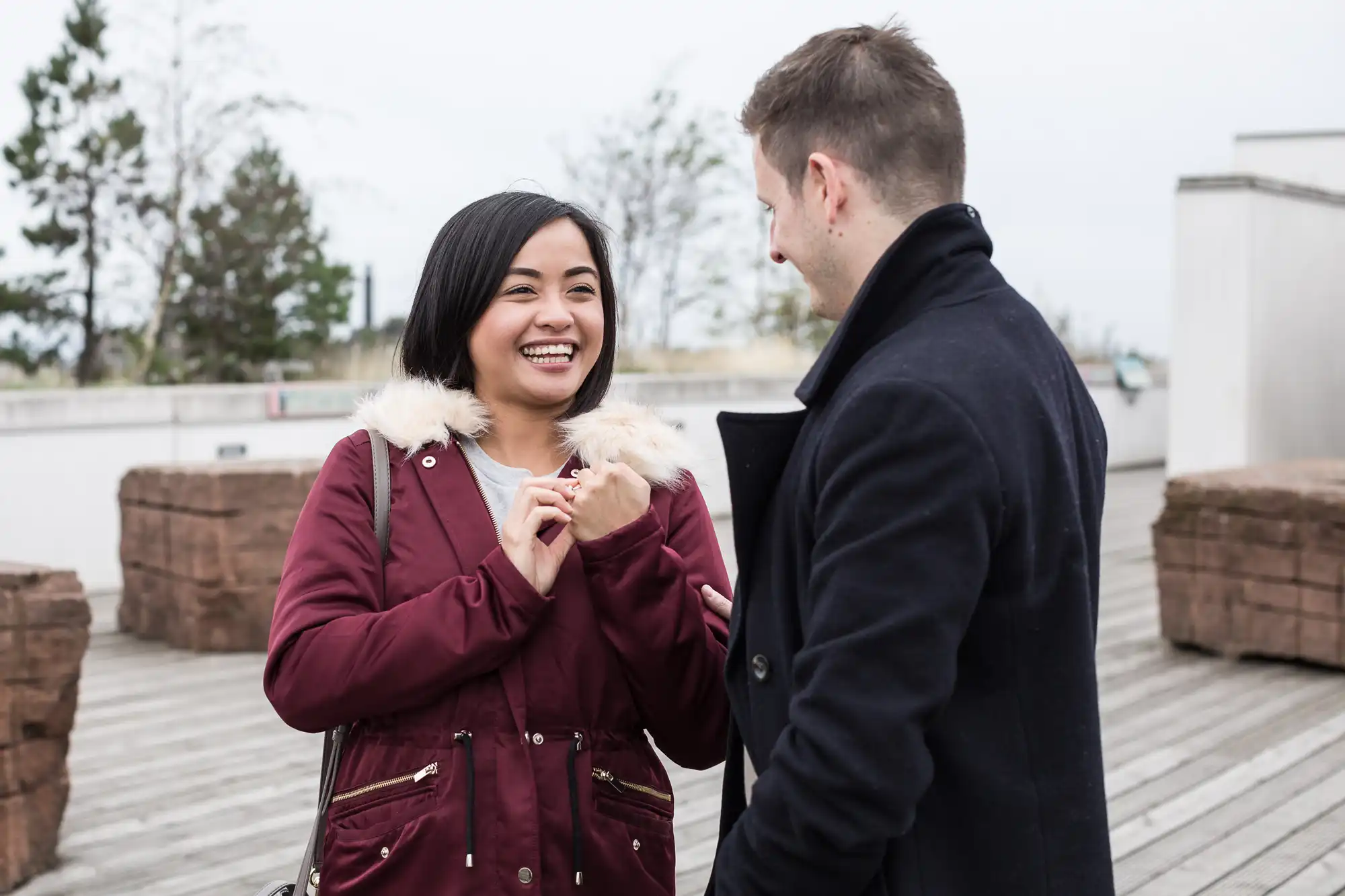 A smiling asian woman in a burgundy jacket talks with a caucasian man in a dark coat on a rooftop.