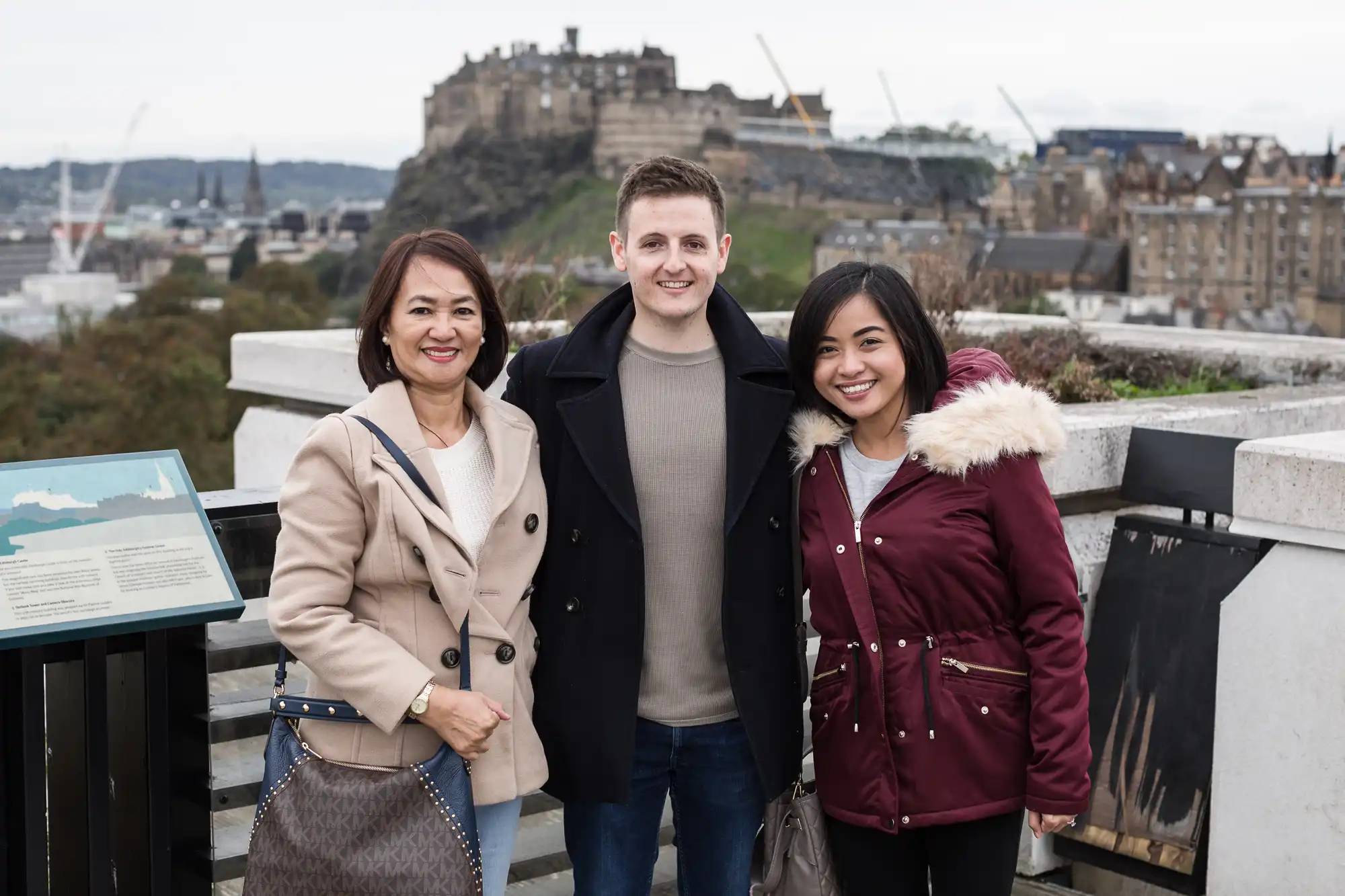Three people smiling at the camera with edinburgh castle in the background. two women and one man stand together, dressed in warm clothing.