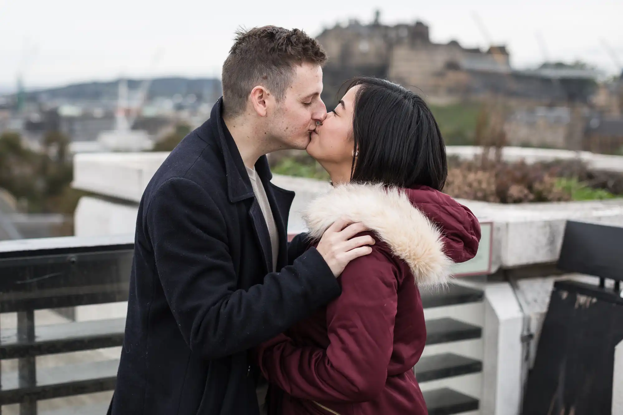 A couple kissing on a rooftop with a blurred cityscape and castle in the background.