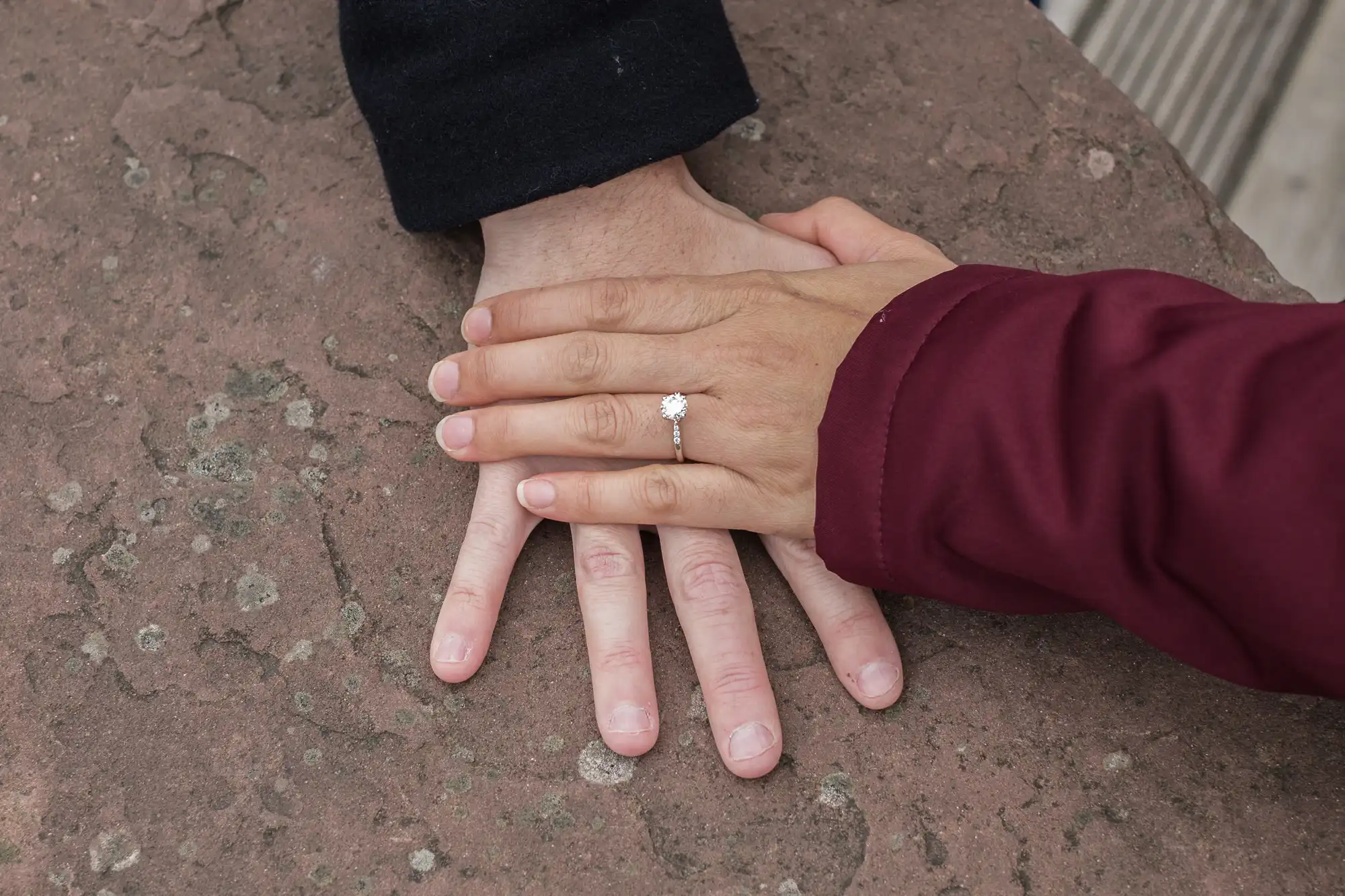 A close-up view of a woman's hand on top of a man's hand, highlighting her engagement ring, resting on a stone surface.