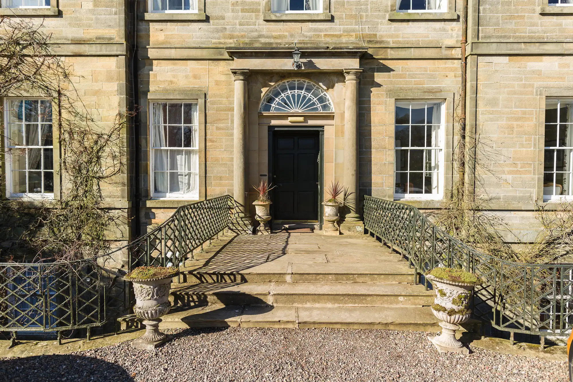 Stone steps lead to a grand black door with a fanlight window, flanked by two columns. The entrance is adorned with potted plants, and symmetrical railings line both sides. Large sash windows and creeping vines are on the light stone façade.
