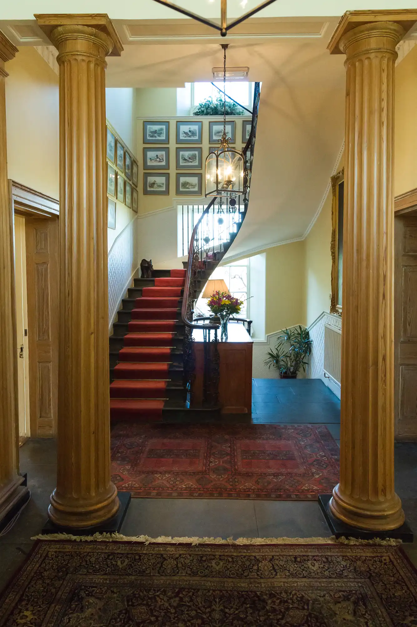 Grand entrance hall with two large wooden columns, red and patterned rugs, a sweeping staircase with red carpet, ornate black balustrade, a chandelier, and framed pictures on the back wall. A bouquet of flowers sits on a small table by the stairs.