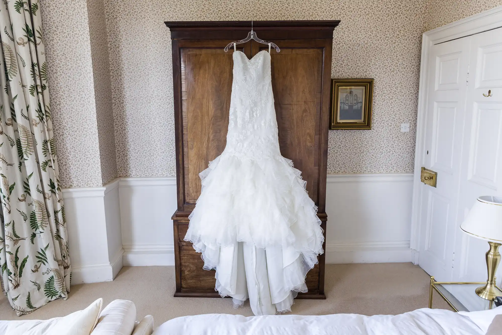 A white, strapless, lace wedding dress with a ruffled tulle skirt hangs on a hanger from a wooden wardrobe in a light, elegant bedroom. The room has cream carpet, patterned wallpaper, and classic decor, including a small framed picture and a lamp.