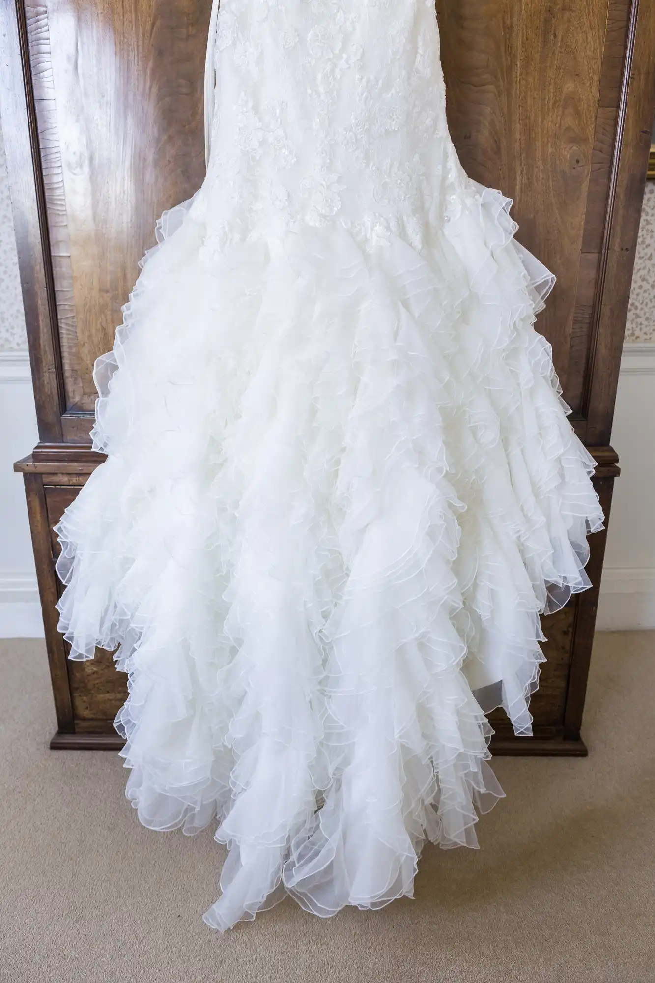 A white wedding dress hangs against a wooden panel. The dress features a fitted, lace-embellished bodice and a voluminous, ruffled skirt made of layers of sheer fabric, giving it a soft and flowing appearance. The lower part spreads out, creating a dramatic look.