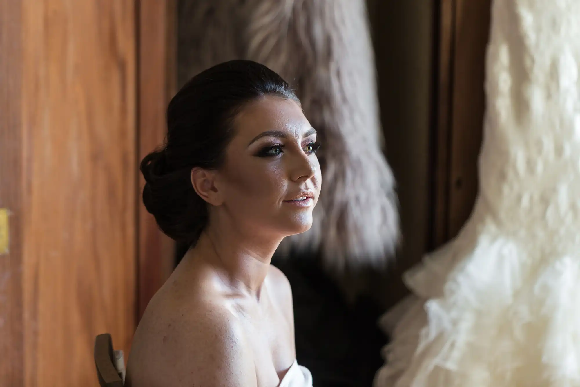 A woman with styled dark hair and subtle make-up, wearing a strapless top, sits in soft natural light. Behind her, a cream-coloured ruffled fabric and textured brown garment hang, creating a warm, elegant atmosphere. She gazes thoughtfully to the side.