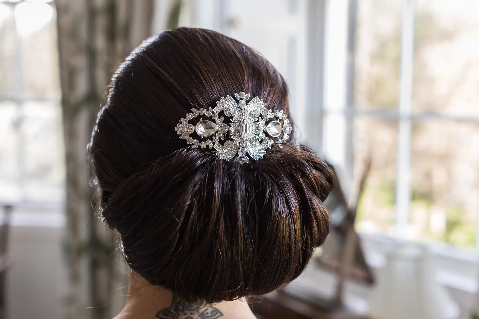 A woman with dark brown hair styled in a low, elegant bun wears a sparkly silver and crystal hair accessory. The accessory features floral and leaf designs. The background is softly blurred, showing large windows and natural light.