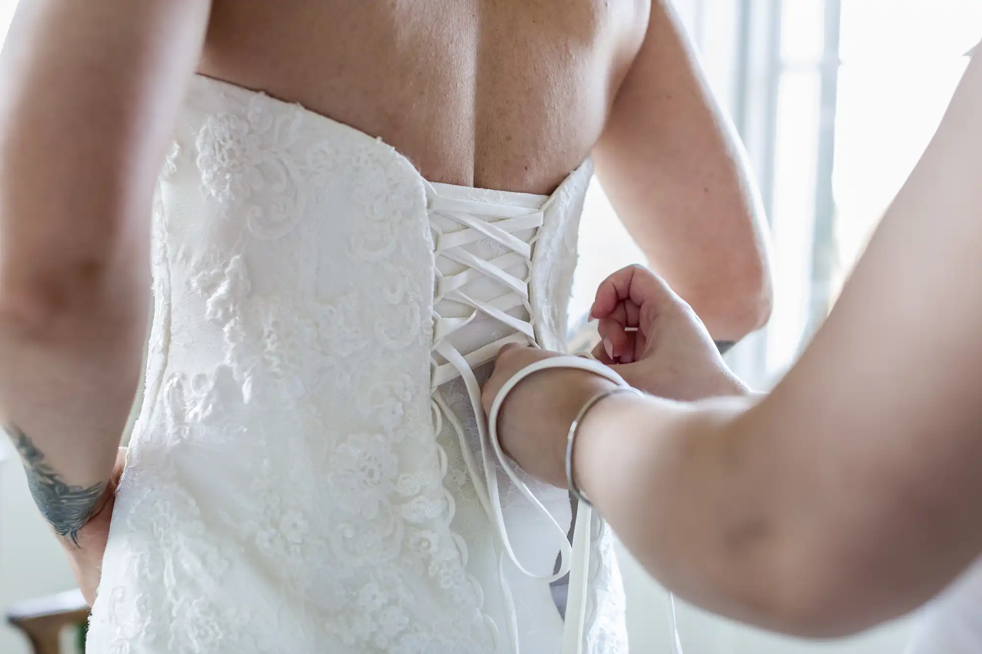 Close-up of a person in a white lace strapless wedding dress being laced up at the back by another person. The second person’s hands are pulling the corset ribbons tight, with light streaming in from a nearby window.