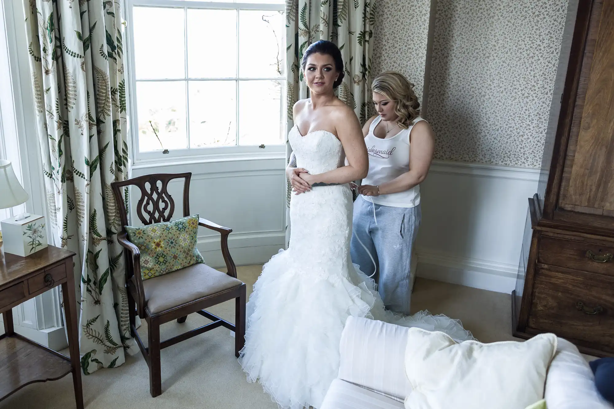 A bride in a strapless white gown stands near a window while another woman in casual clothes fastens the back of her dress. The room is bright with patterned curtains, a wooden chair, and neutral walls, creating a calm, intimate atmosphere.