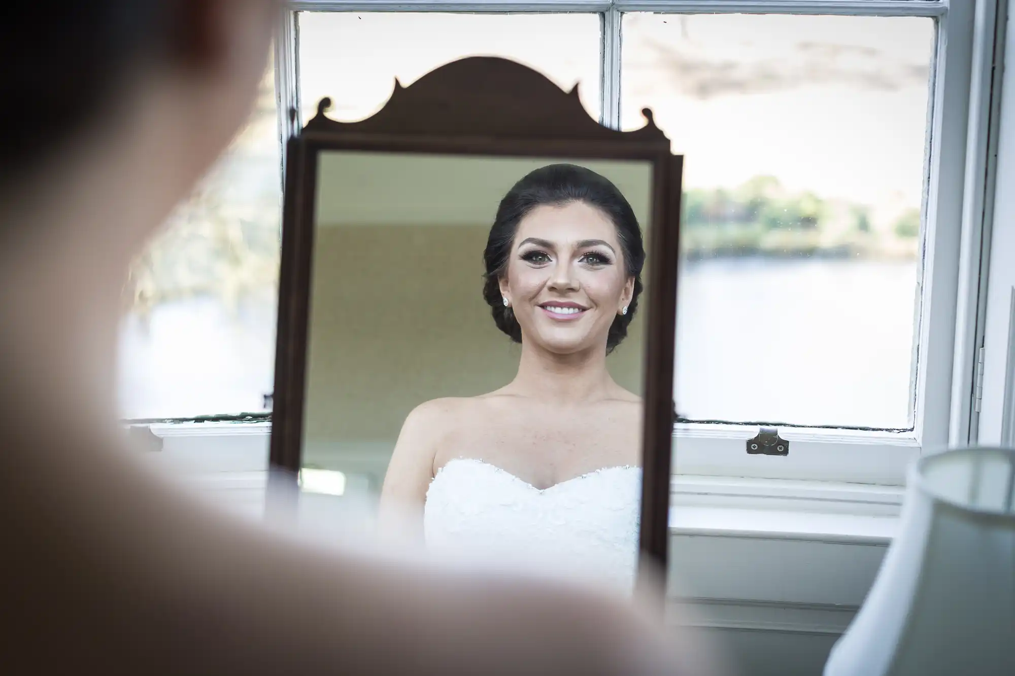 A bride in a strapless white dress is smiling as she looks at her reflection in a vintage mirror. The background shows a bright window with an outdoor view, and a lamp is visible on the right side of the image.