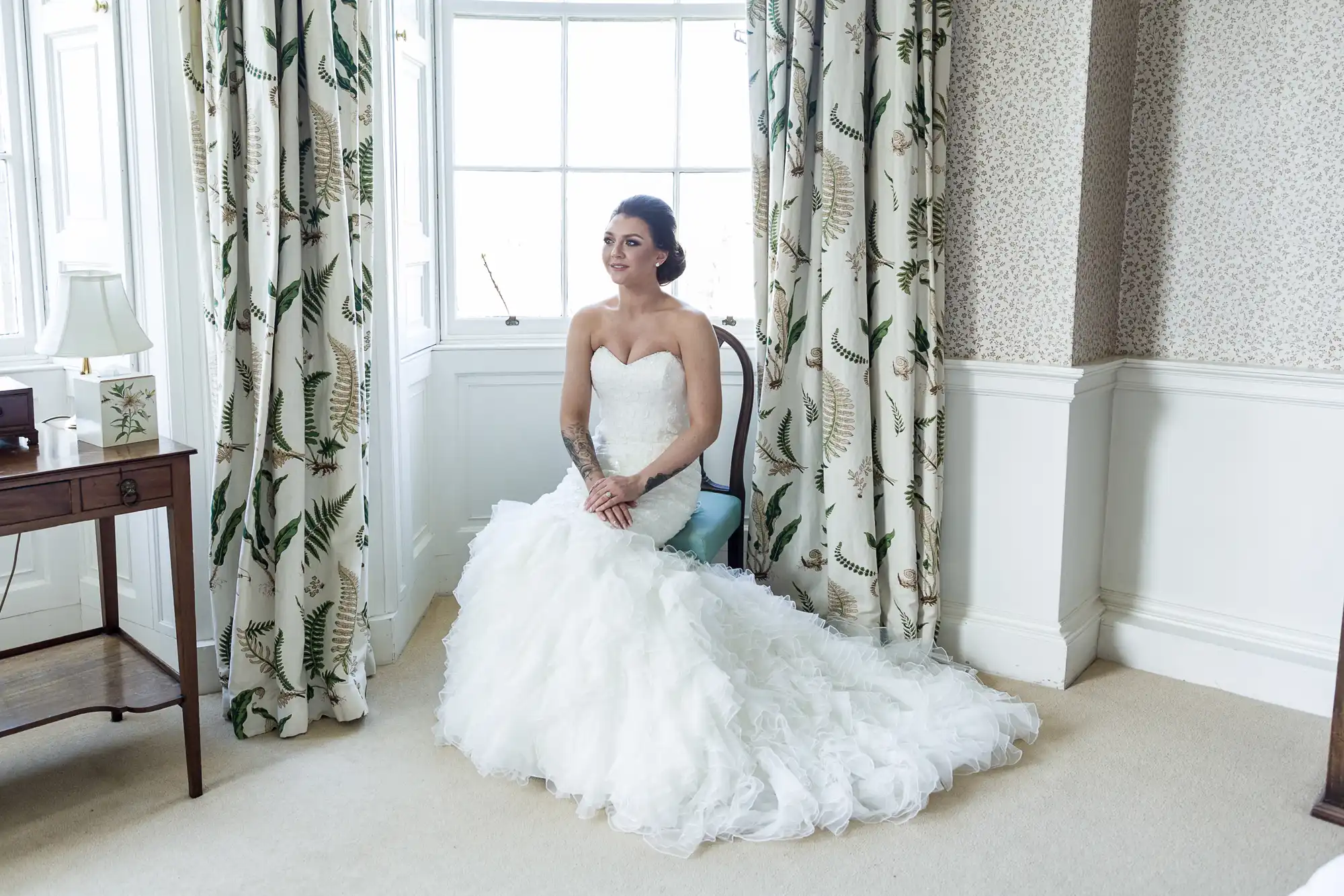 A bride in a strapless white wedding dress with a full ruffled skirt sits on a chair by a large window in a bright, elegant room with patterned curtains, a small desk, and cream carpet. She smiles softly, her hair styled up.