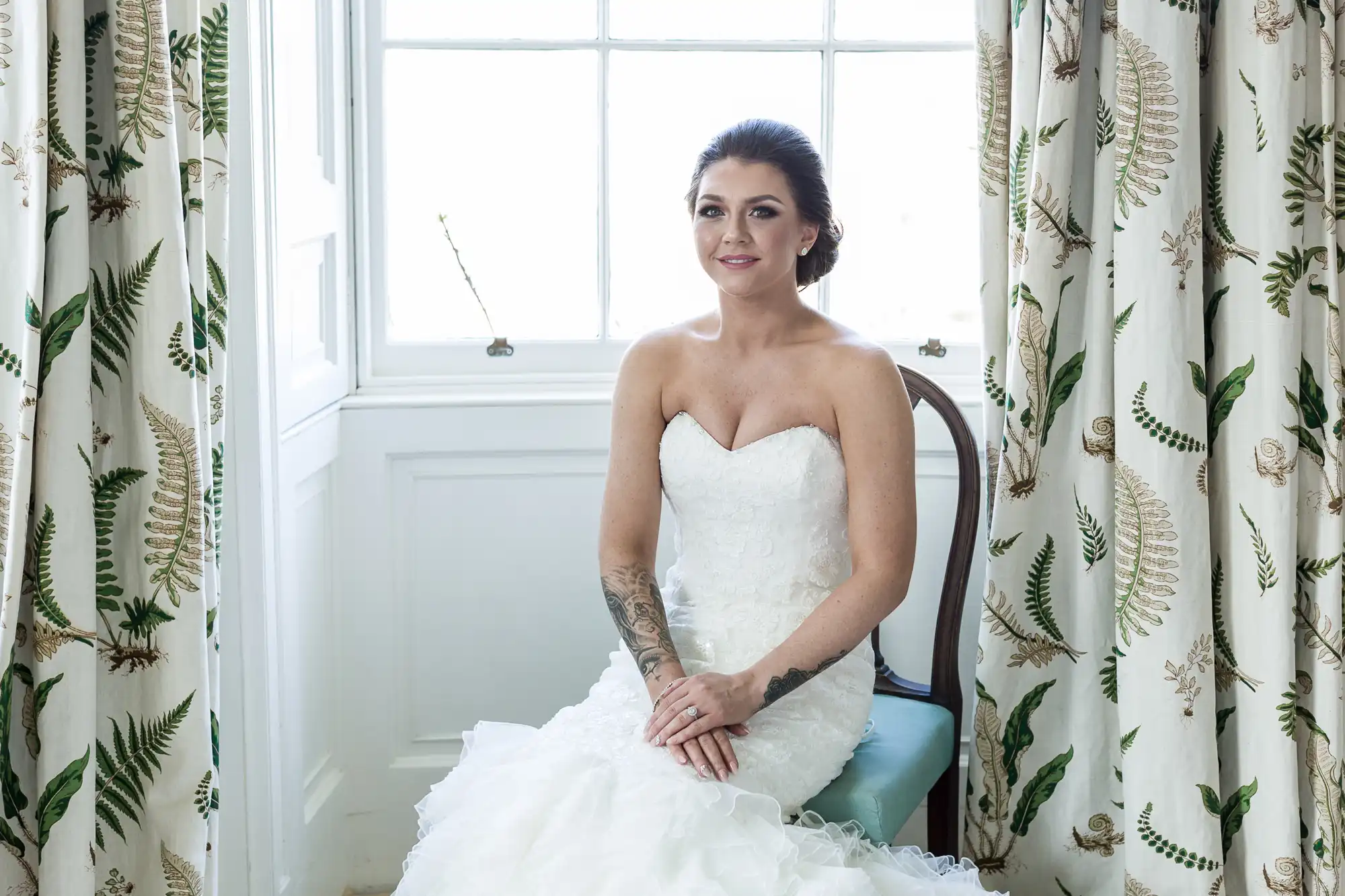 A bride with dark hair in an updo sits on a chair in front of a bright window, wearing a strapless white lace wedding dress. She has visible tattoos on her arms. The room has white walls and curtains with fern patterns. She smiles softly.