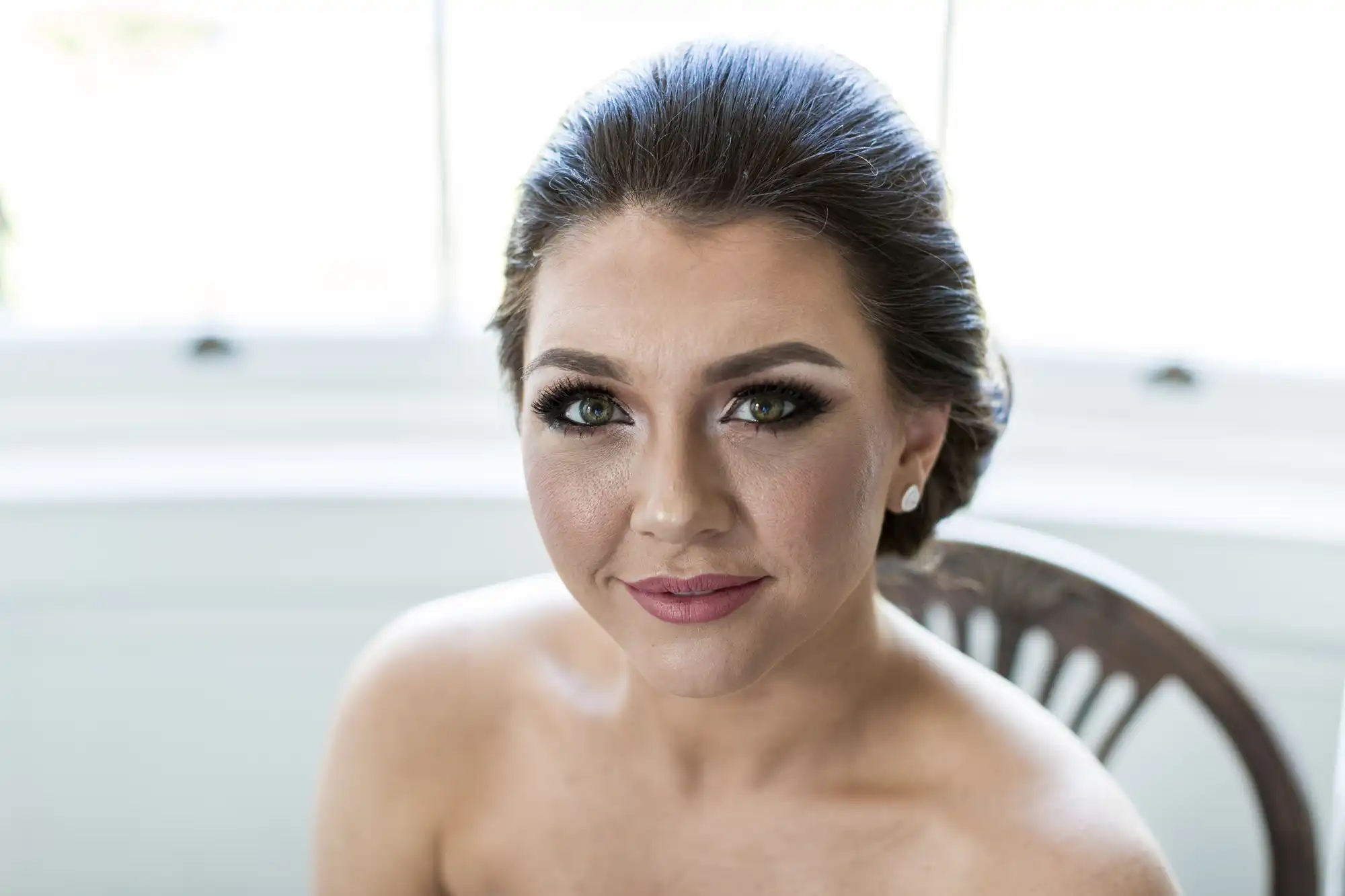 A woman with styled dark hair sits in front of a bright window. She has dramatic eye make-up, long lashes, rosy lips, and pearl earrings. She looks directly at the camera, smiling softly, with bare shoulders visible, suggesting formal or bridal attire.