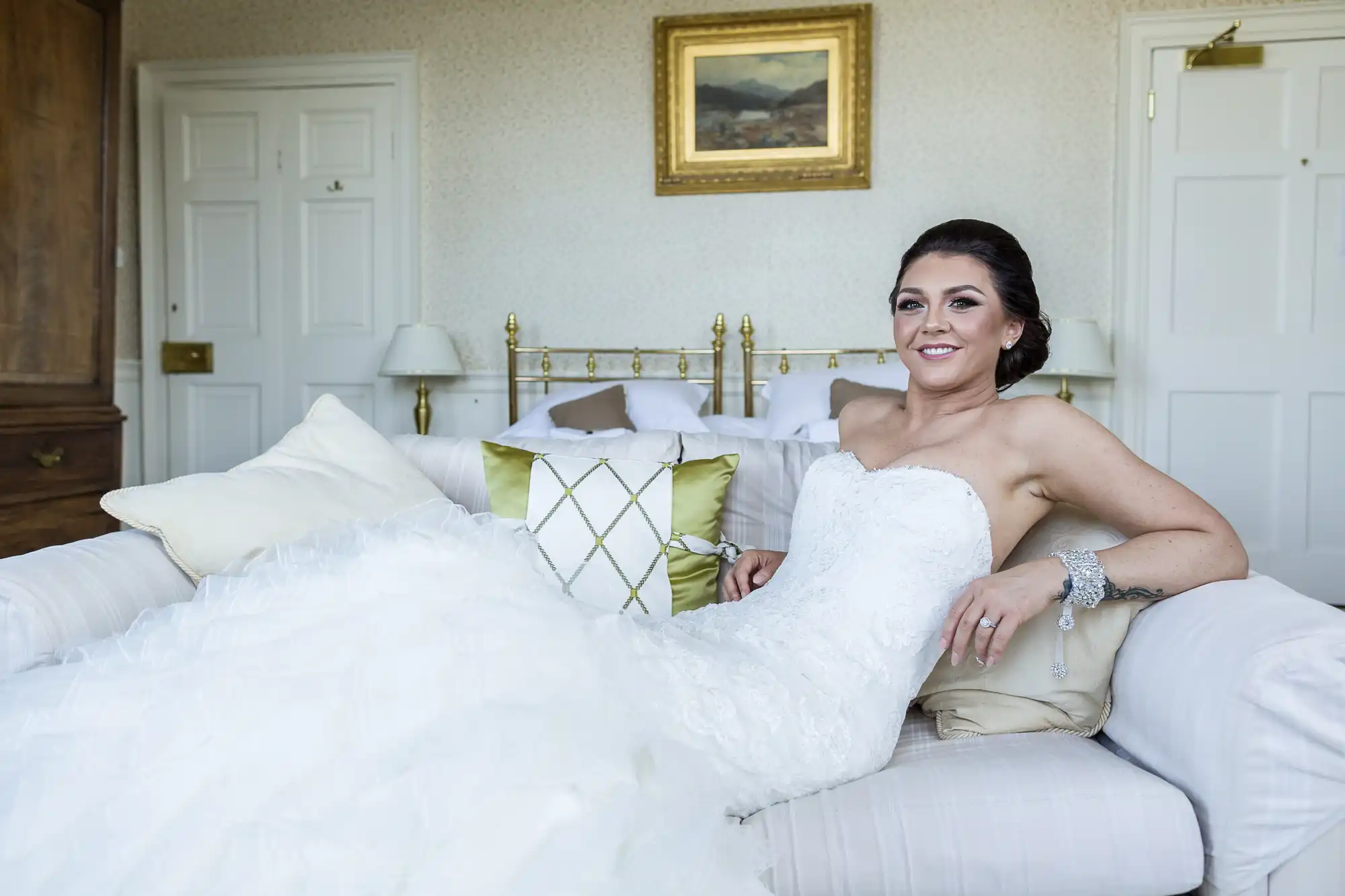 A bride in a strapless white wedding dress reclines on a cream sofa with decorative cushions, smiling at the camera. Behind her is a neatly made bed, two doors, and a framed landscape painting on the wall in a softly lit, elegant room.