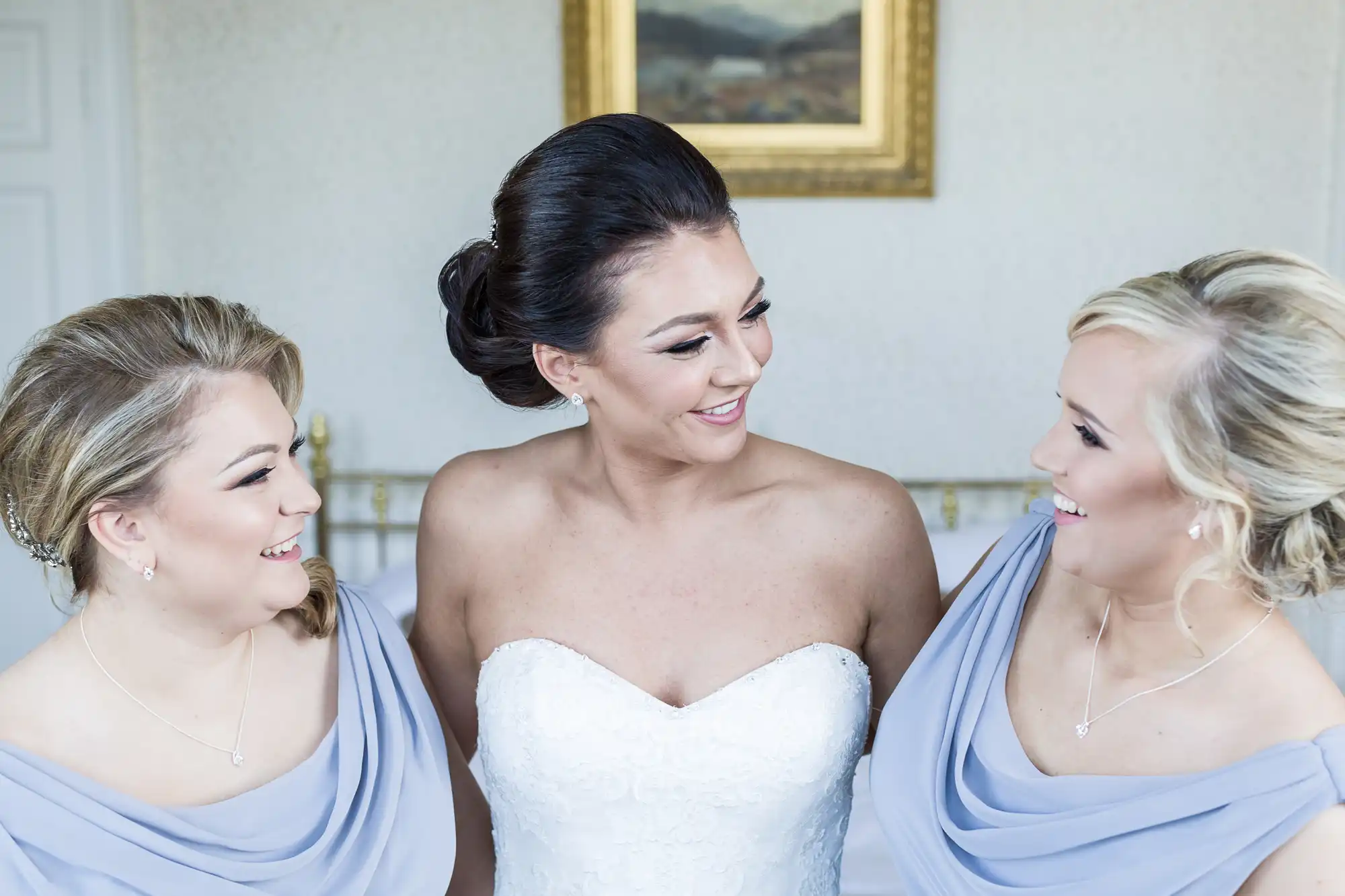 Three women smile at each other. The woman in the centre wears a white strapless wedding dress, while the two on either side wear matching light lavender dresses. All have styled hair and necklaces. A framed picture hangs on a wall behind them.