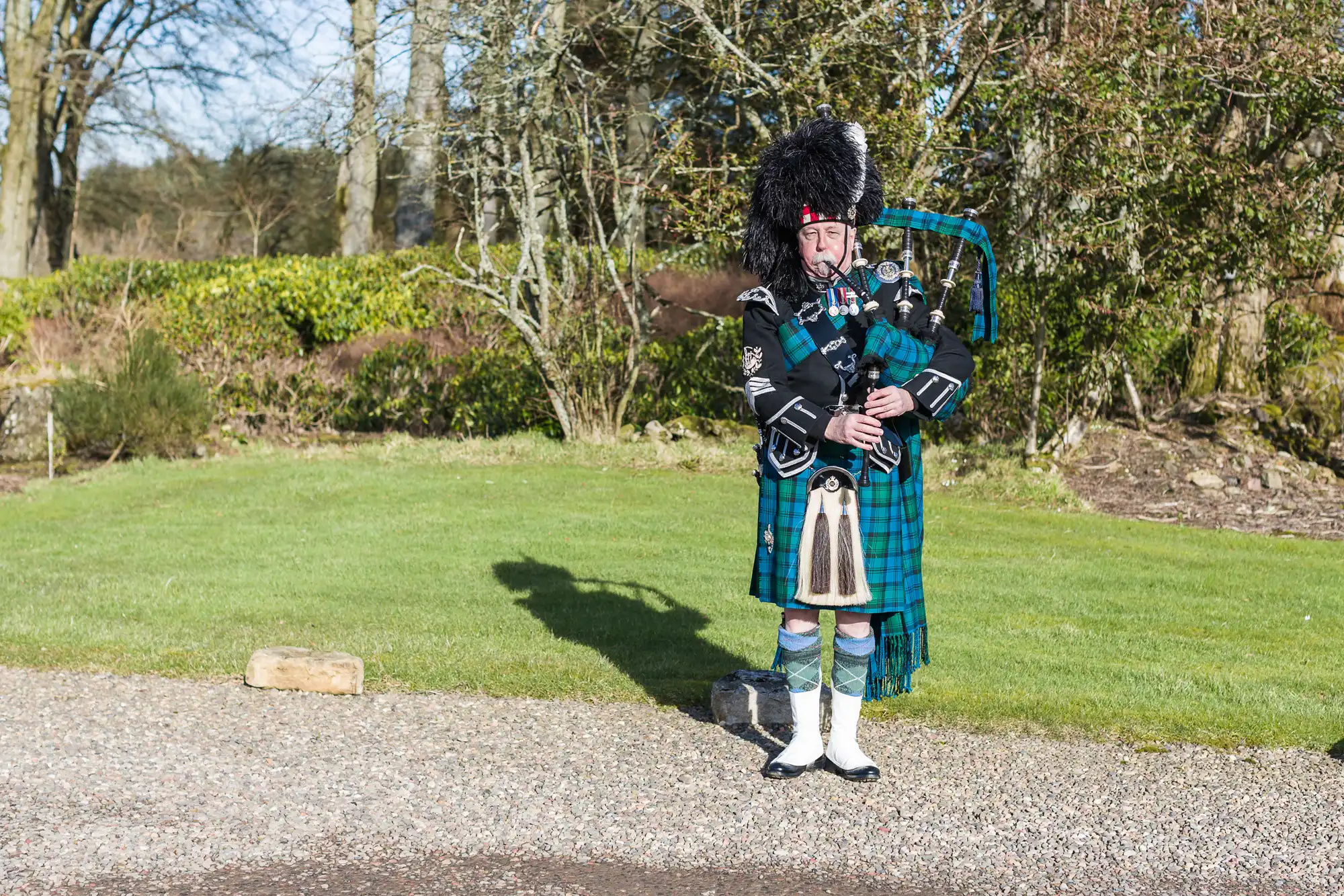 A man in traditional Scottish Highland dress, including a feathered hat and tartan kilt, plays bagpipes outdoors on a sunny day. He stands on gravel near a green lawn with trees and shrubs in the background.