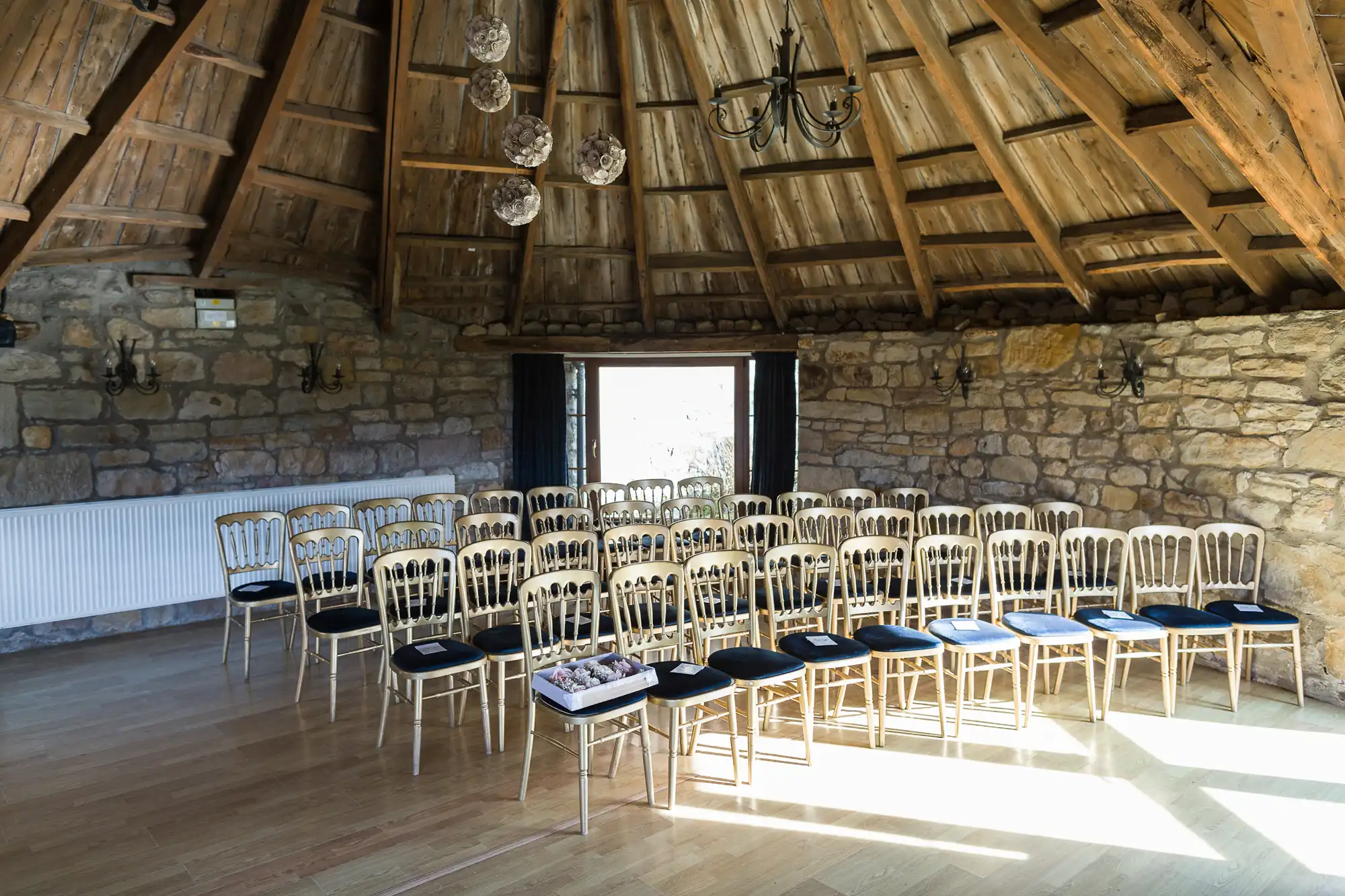 A rustic room with stone walls and a wooden beam ceiling. Rows of gold chairs with black cushions face a window at the back. Sunlight streams in, illuminating the wooden floor. Chandeliers and wall sconces add charm to the cosy, intimate setting.