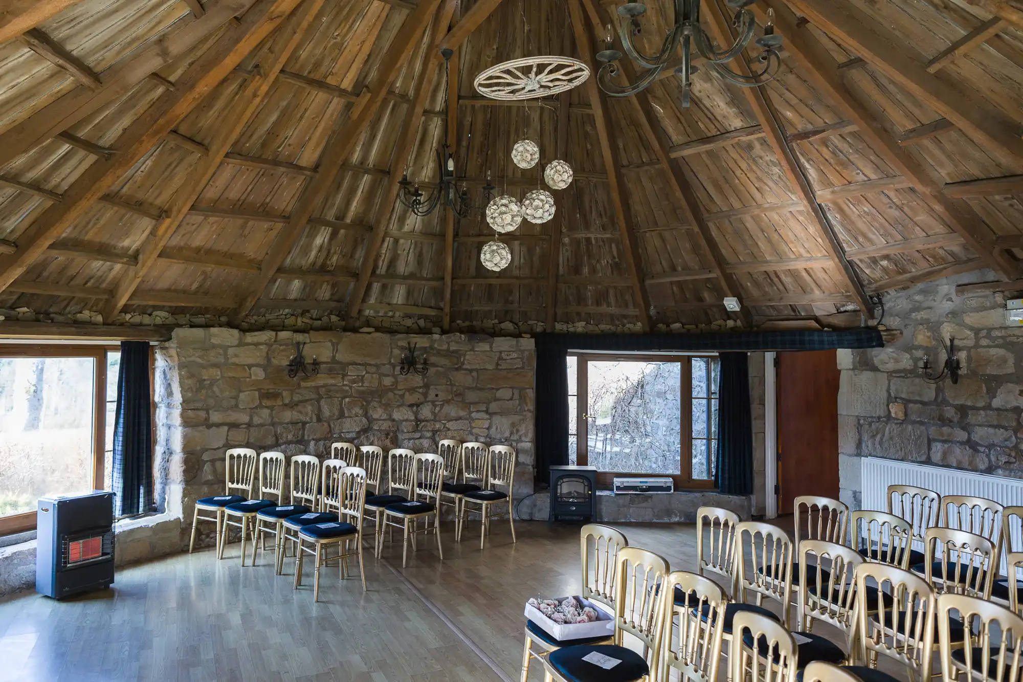 A rustic circular room with a high wooden-beam ceiling, stone walls, and large windows. Gold chairs with dark cushions are arranged in rows, facing a small fireplace and a window. A chandelier and wagon wheel hang from the ceiling.