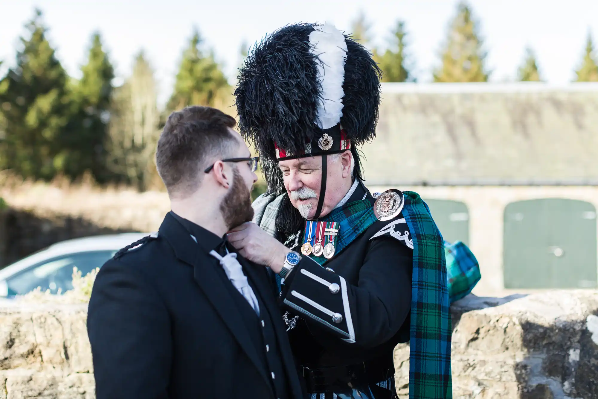 Two men in formal Scottish attire stand outside. One, wearing a feathered hat, medals, and a tartan sash, adjusts the other man's collar. Both appear focused on the task. Trees and stone buildings are visible in the bright background.