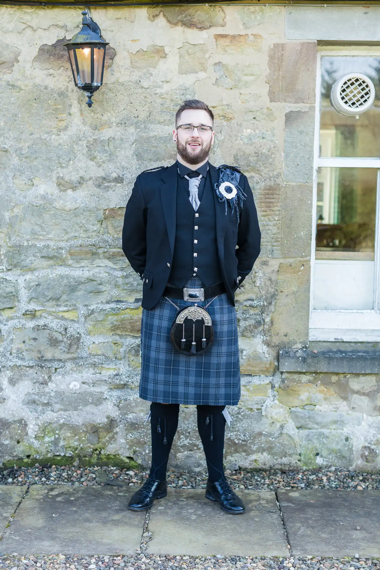 A man stands in front of a stone wall, wearing traditional Scottish attire: a black jacket, waistcoat, and a blue-and-grey tartan kilt with a sporran. He has glasses, a beard, and a buttonhole. There’s a window and lantern on the wall behind him.