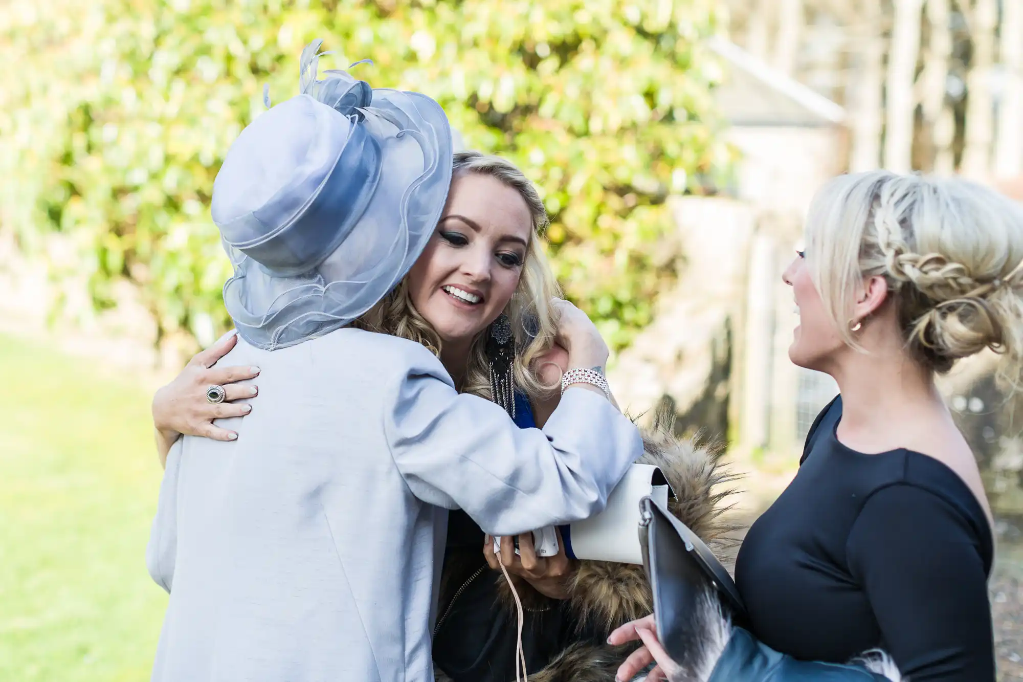 Three women greet each other outdoors. One woman in a light blue hat and coat hugs a smiling woman with long blonde hair. Another woman with blonde hair, in a black dress, stands nearby, laughing and holding a clutch bag. Greenery and sunlight are in the background.