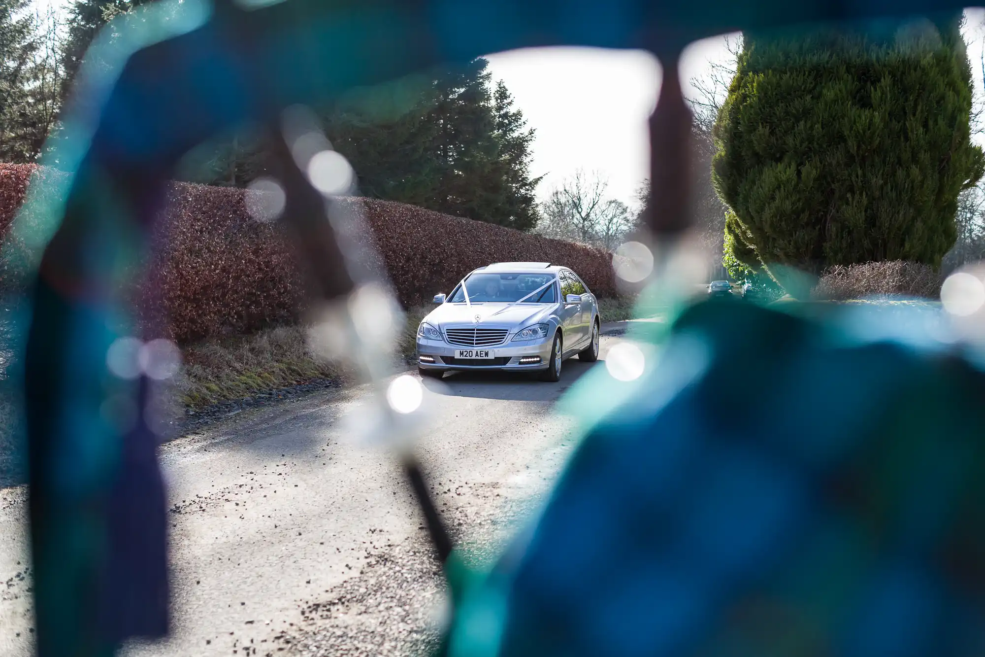 A silver car is parked on a quiet road lined with leafless bushes and trees. The photo is taken from behind a partially blurred, blue-green object, framing the car in the centre of the image. Sunlight casts shadows across the scene.