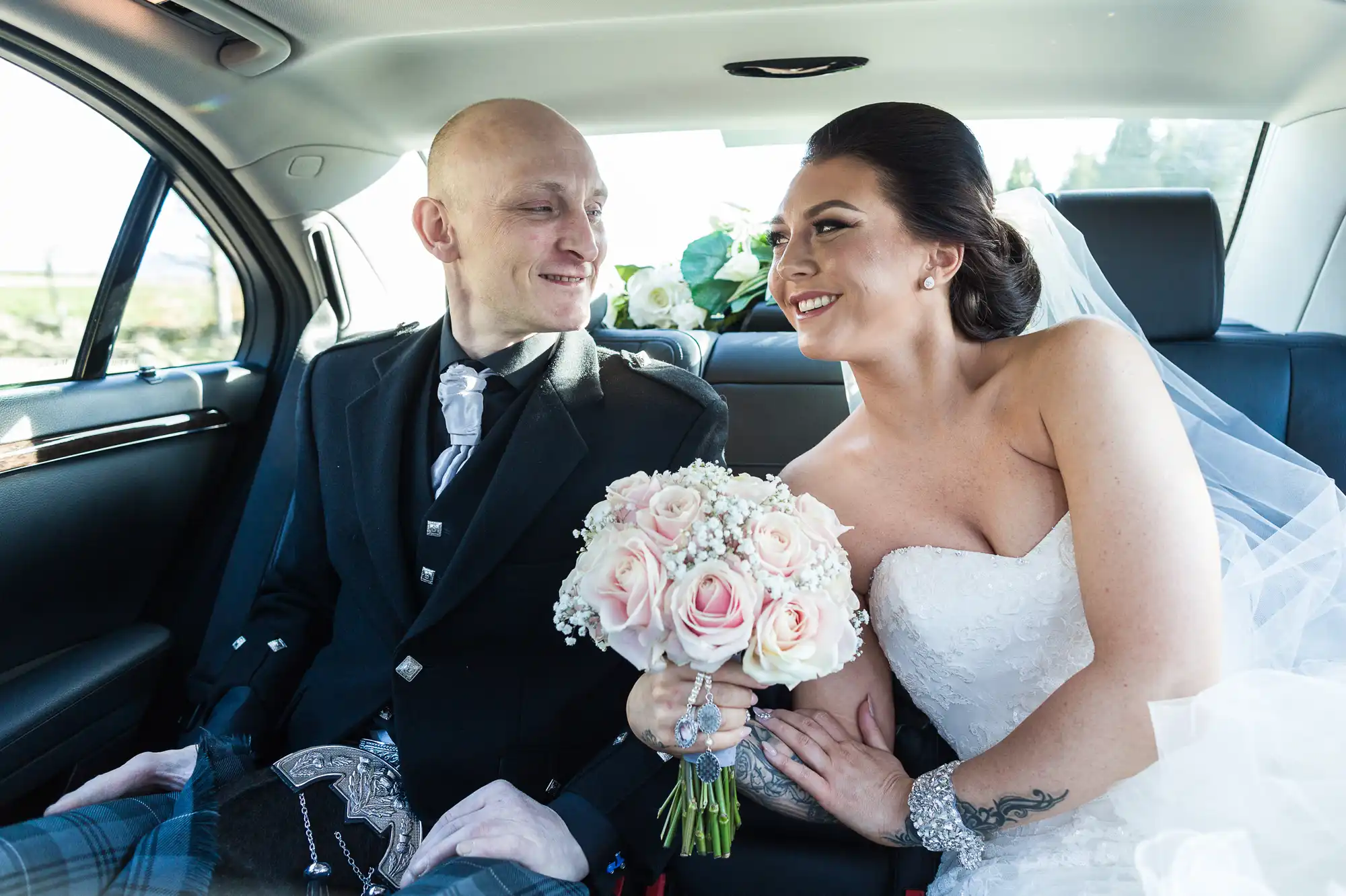 A bride in a white strapless gown holding a bouquet of light pink and white roses smiles lovingly at a groom wearing a black jacket, kilt, and tie. They sit close together in the backseat of a car, holding hands and looking into each other's eyes.