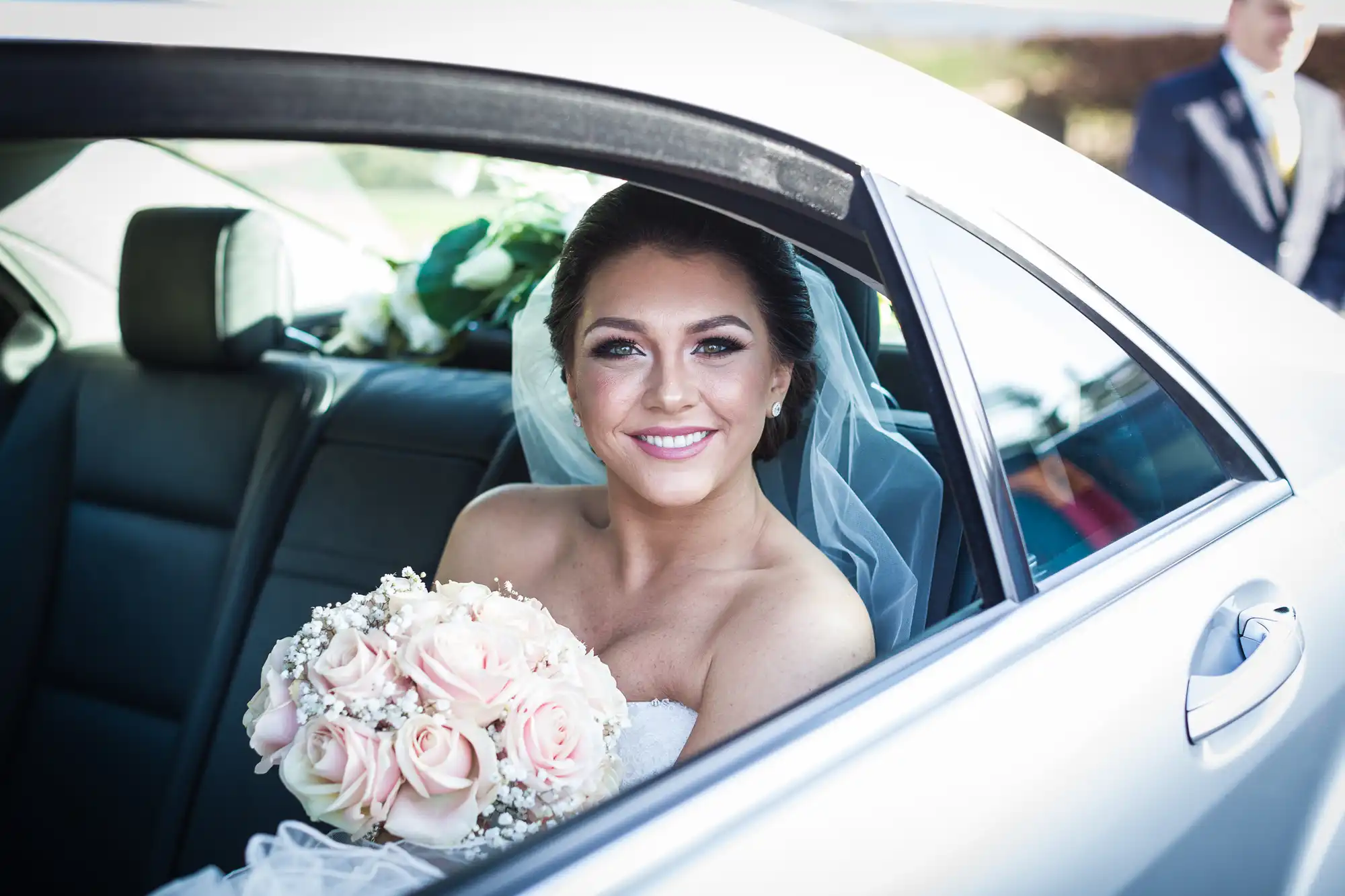 A smiling bride in a strapless white dress and veil sits in the backseat of a car, holding a bouquet of pale pink roses and baby's breath. She looks out the open window, ready for her wedding, with soft sunlight illuminating her face.
