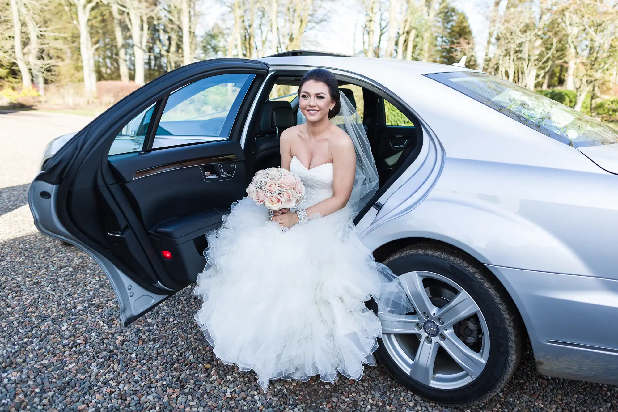 A smiling bride in a white wedding dress and veil holds a bunch of pale flowers while sitting in the back seat of a silver car with the door open, on a gravel drive surrounded by bare trees.