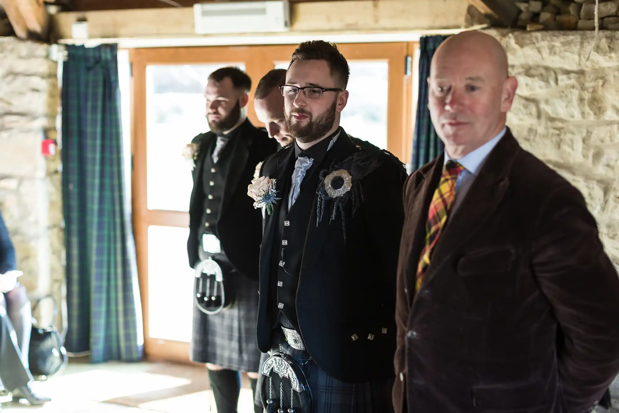 Three men stand indoors near a stone wall and large window with blue curtains. Two men wear traditional Scottish kilts and jackets with floral buttonholes, while the third man in front wears a brown jacket and checked tie, looking towards the camera.