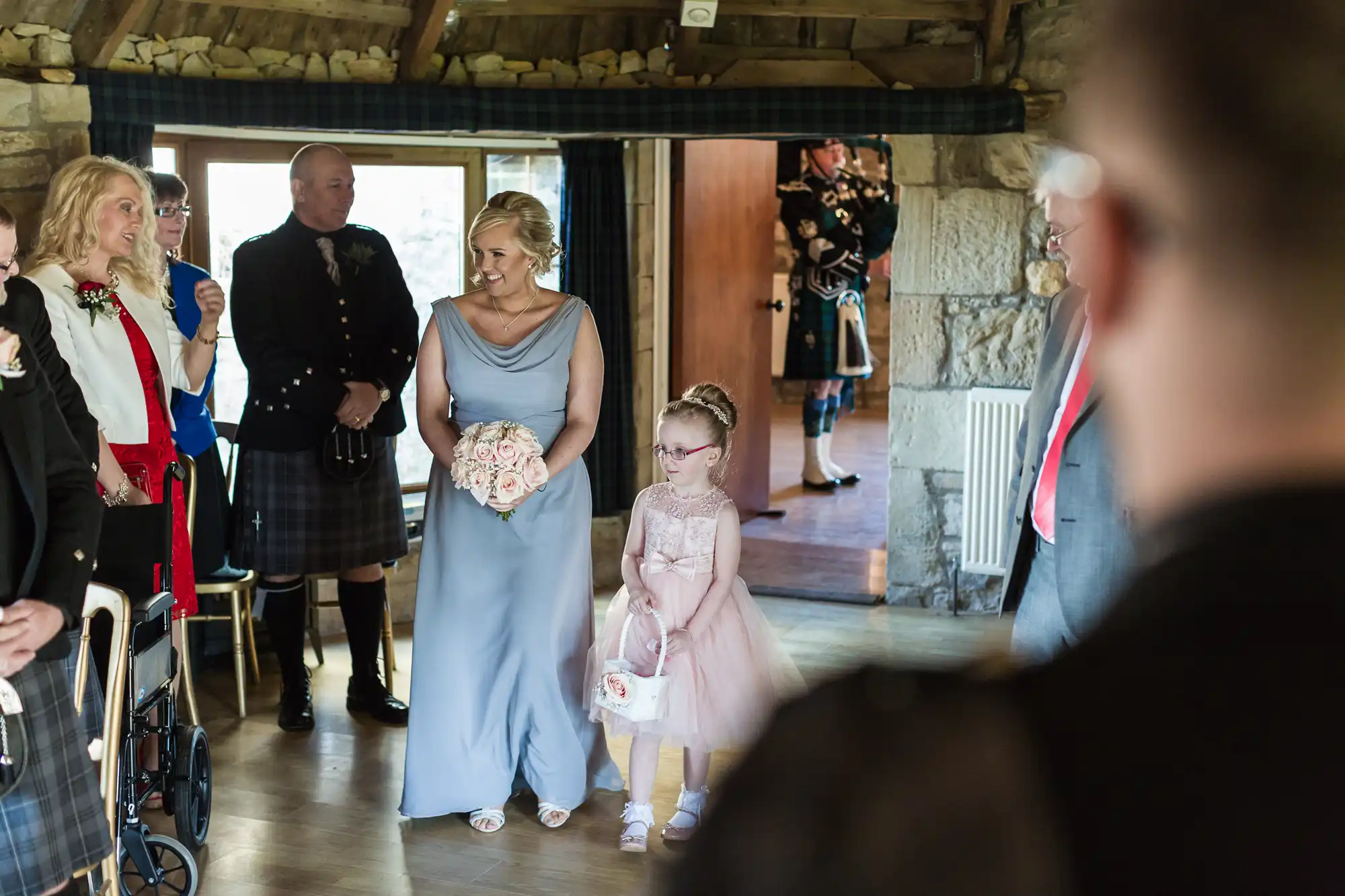 A smiling woman in a pale blue gown holds a bouquet, walking with a young girl in a pink dress and glasses. Guests in semi-formal attire watch them inside a rustic stone venue. A bagpiper in tartan is visible in the background near the doorway.