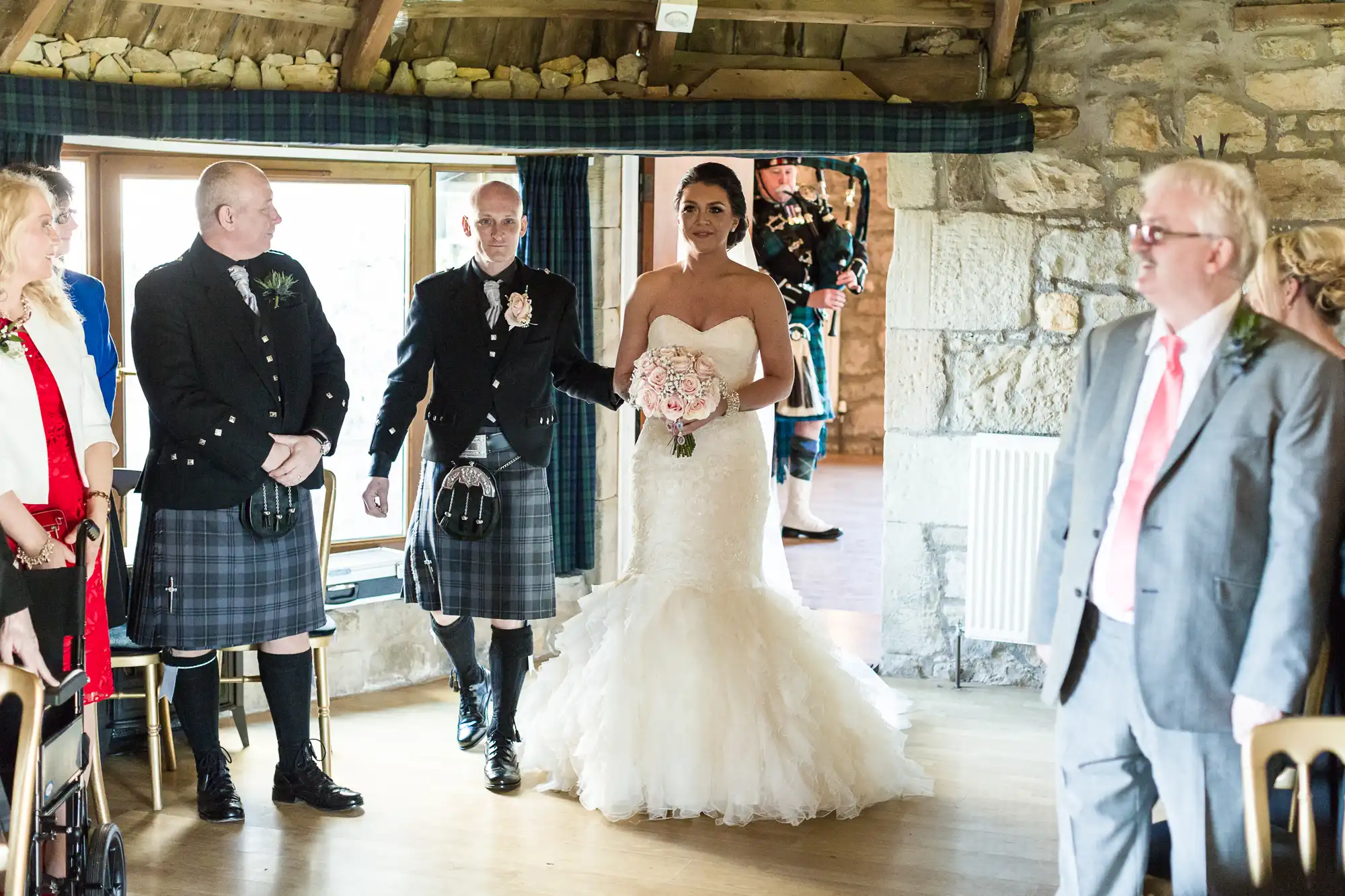 A bride in a strapless white gown holds a bouquet and walks down the aisle with two men in Scottish kilts. Guests, some in formal attire, watch. The setting is a rustic stone room with wooden beams and sunlight streaming through a window.