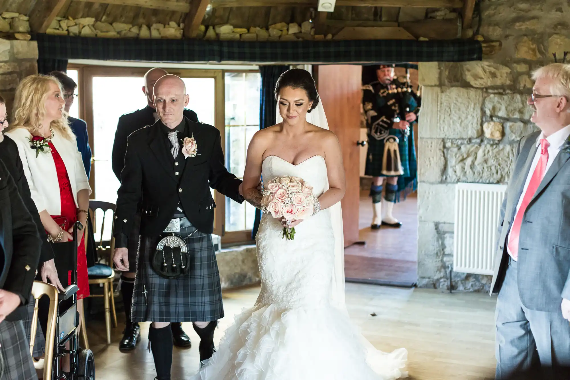 A bride in a strapless white dress holds a bouquet, walking down the aisle with an older man in a kilt. Guests are seated on either side, and two bagpipers in traditional attire stand in the doorway of a rustic stone-walled venue.