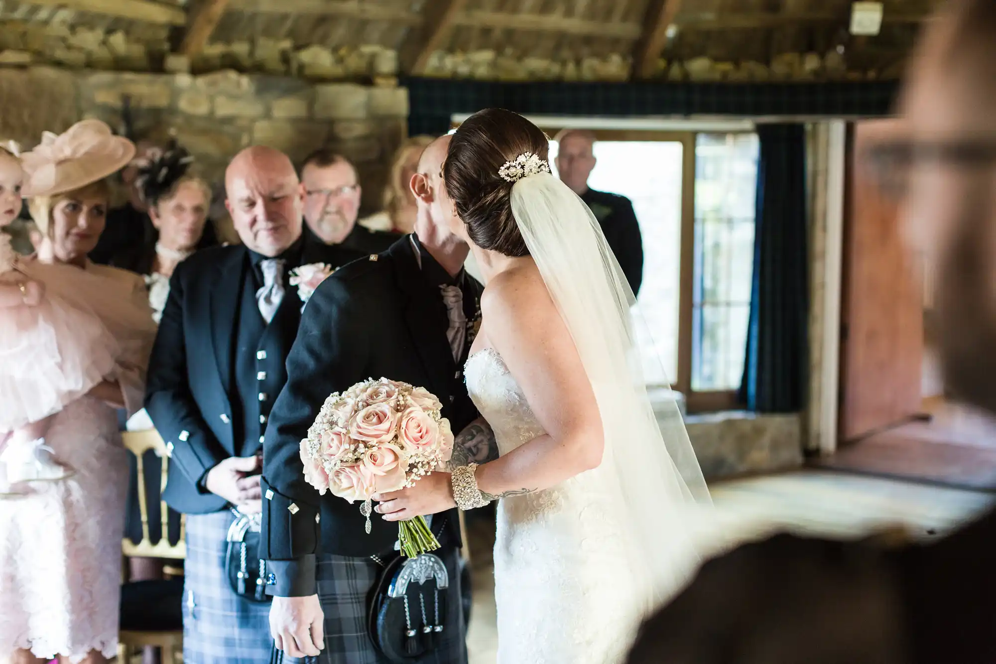 A bride in a white dress with a veil and jewelled hairpiece holds a pink rose bouquet and kisses a man in a kilt at an indoor wedding ceremony, while guests in formal attire watch with emotional expressions.