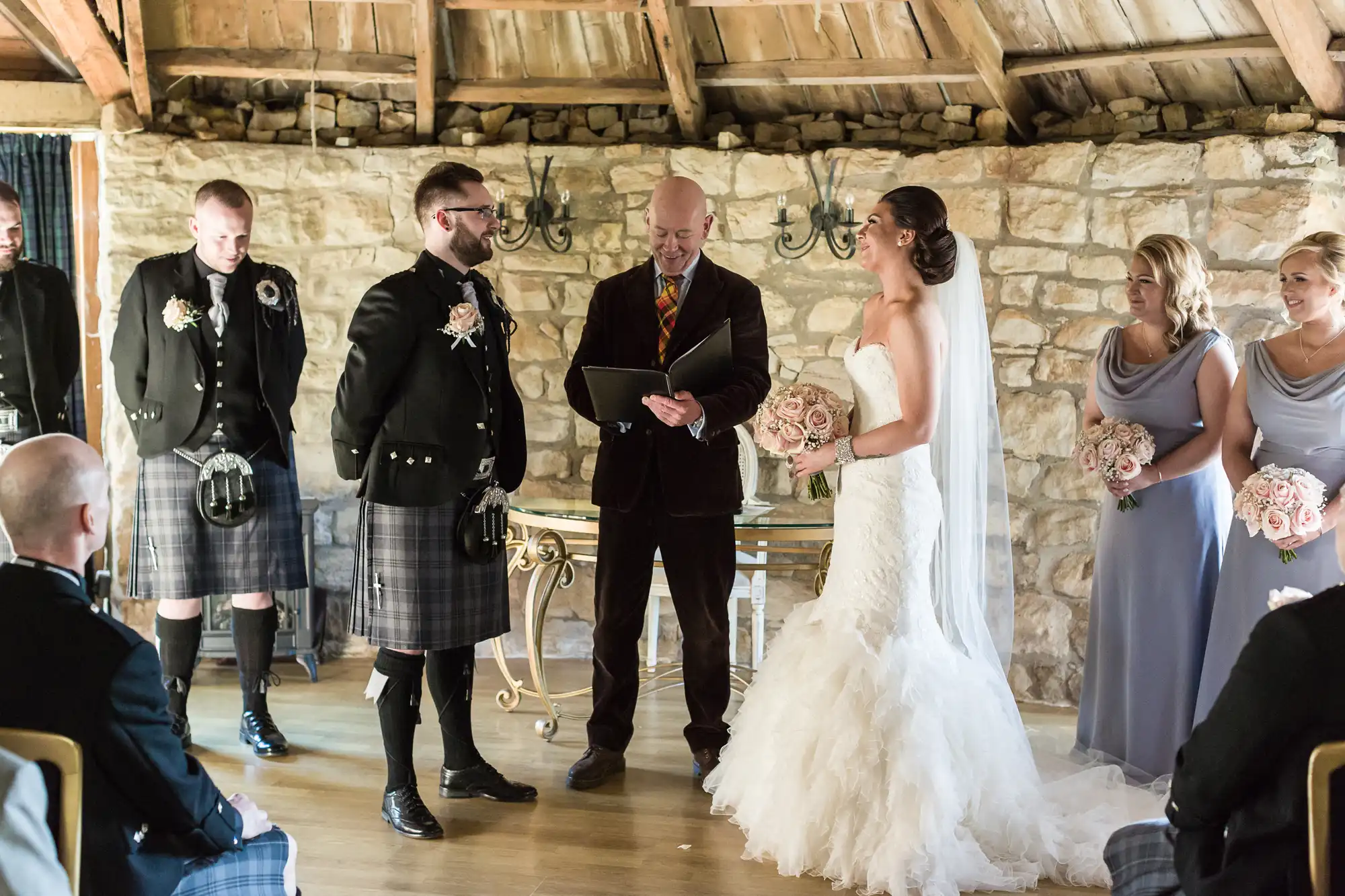 A bride in a white strapless gown and veil holds a bouquet, facing the groom in a kilt and jacket. They stand before an officiant in a rustic room with stone walls and wooden beams, surrounded by attendants in kilts and lavender dresses.