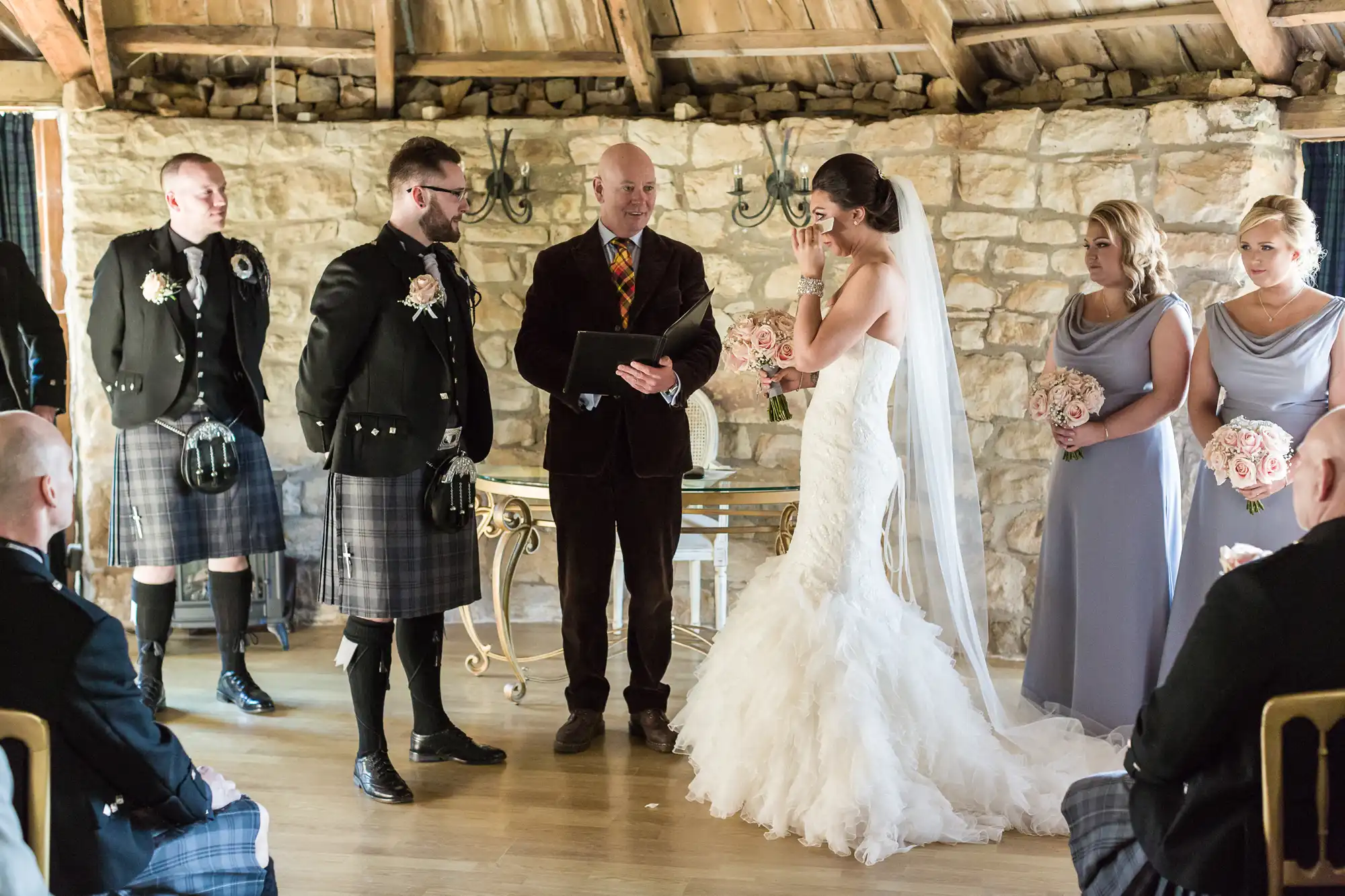 A bride in a white dress wipes her eyes during a rustic indoor wedding ceremony. The groom and officiant stand to her left, both in kilts. Three bridesmaids in light grey dresses hold bouquets to her right. Guests are seated in the foreground. Stone walls and wooden beams surround them.