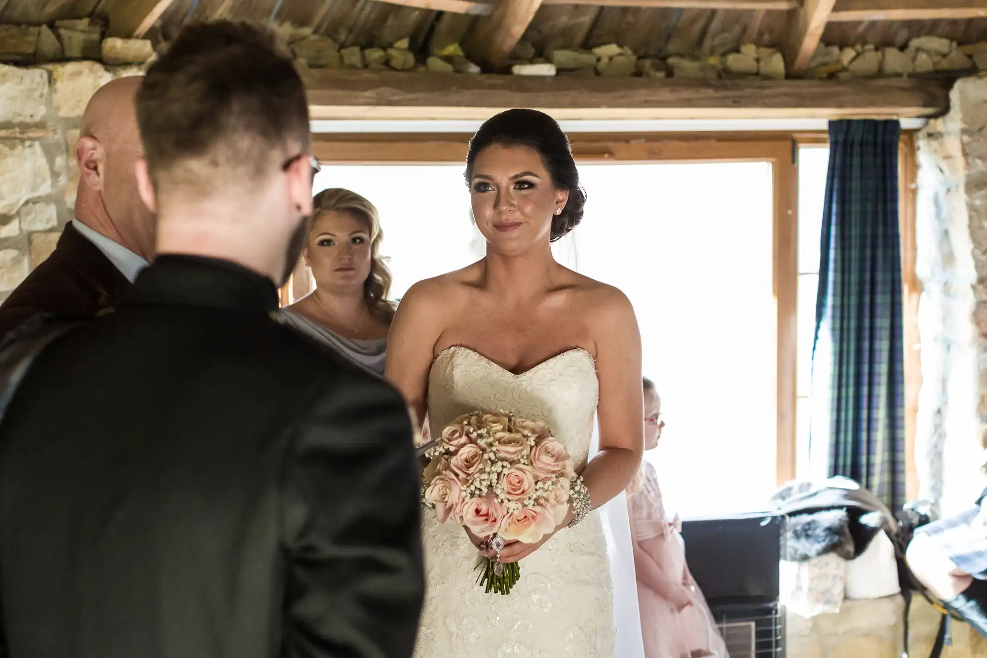 A bride in a strapless white gown holds a bouquet of pale pink roses, smiling at a man in a black suit. Two bridesmaids stand behind her inside a rustic stone venue with exposed wooden beams and natural light from a large window.