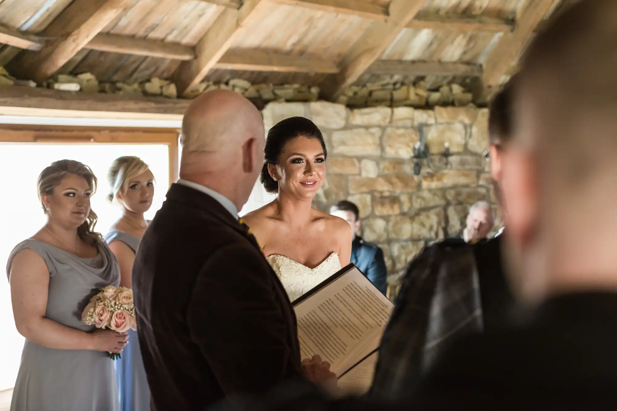 A bride in a white dress smiles during a rustic indoor wedding ceremony. She faces a bald officiant holding a book. Bridesmaids in light purple dresses with bouquets stand nearby, whilst guests watch in a stone-walled room with wooden beams.