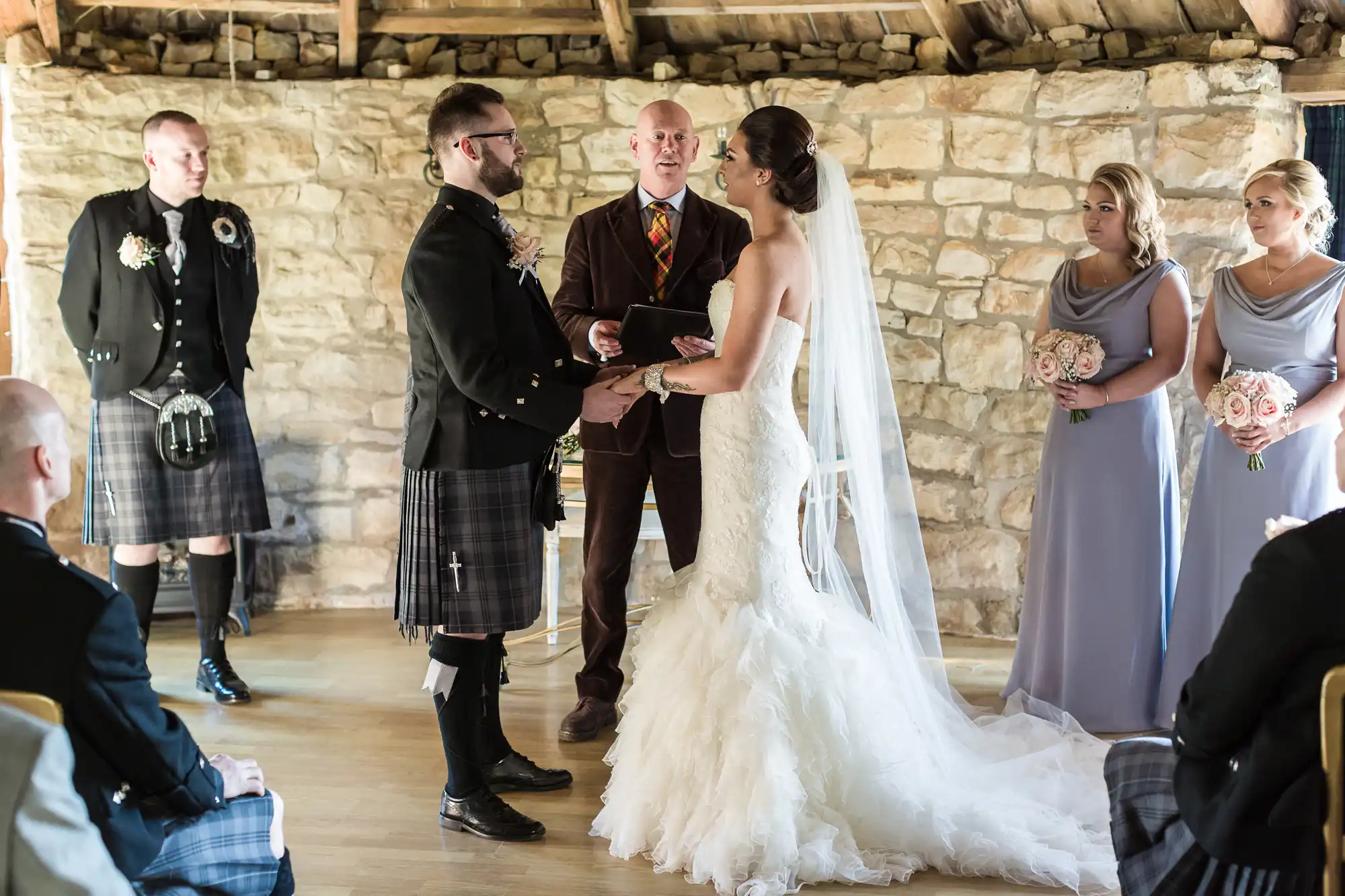 A bride in a strapless white gown and veil holds hands with a groom in a black jacket and kilt during a wedding ceremony. An officiant stands in the centre, with bridesmaids in lavender dresses and groomsmen in kilts nearby, all inside a rustic stone room.