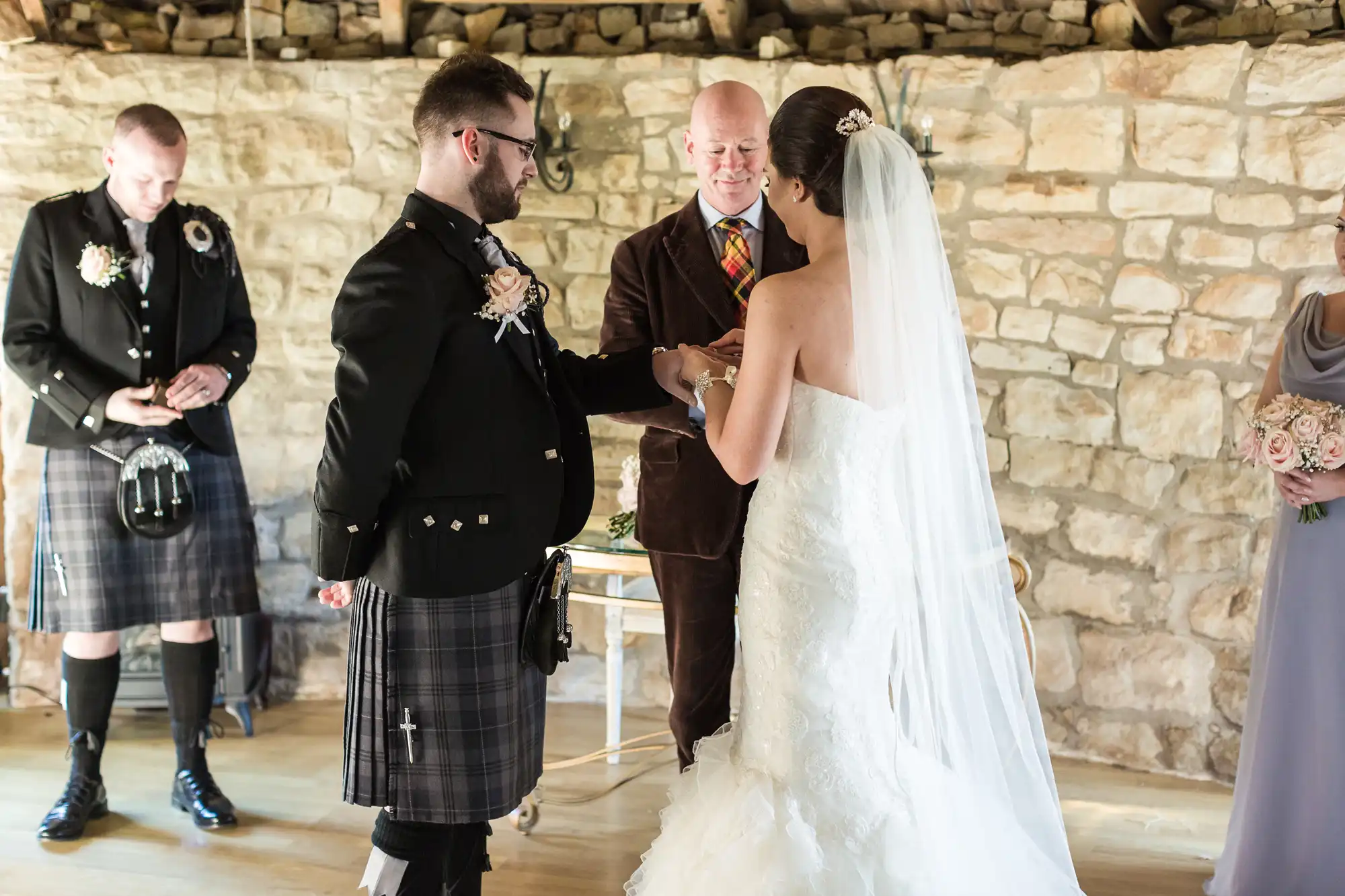 A bride in a white dress and veil holds hands with a groom in a black jacket and kilt. An officiant stands behind them, and attendants in formal attire are nearby. The ceremony takes place indoors against a stone wall.
